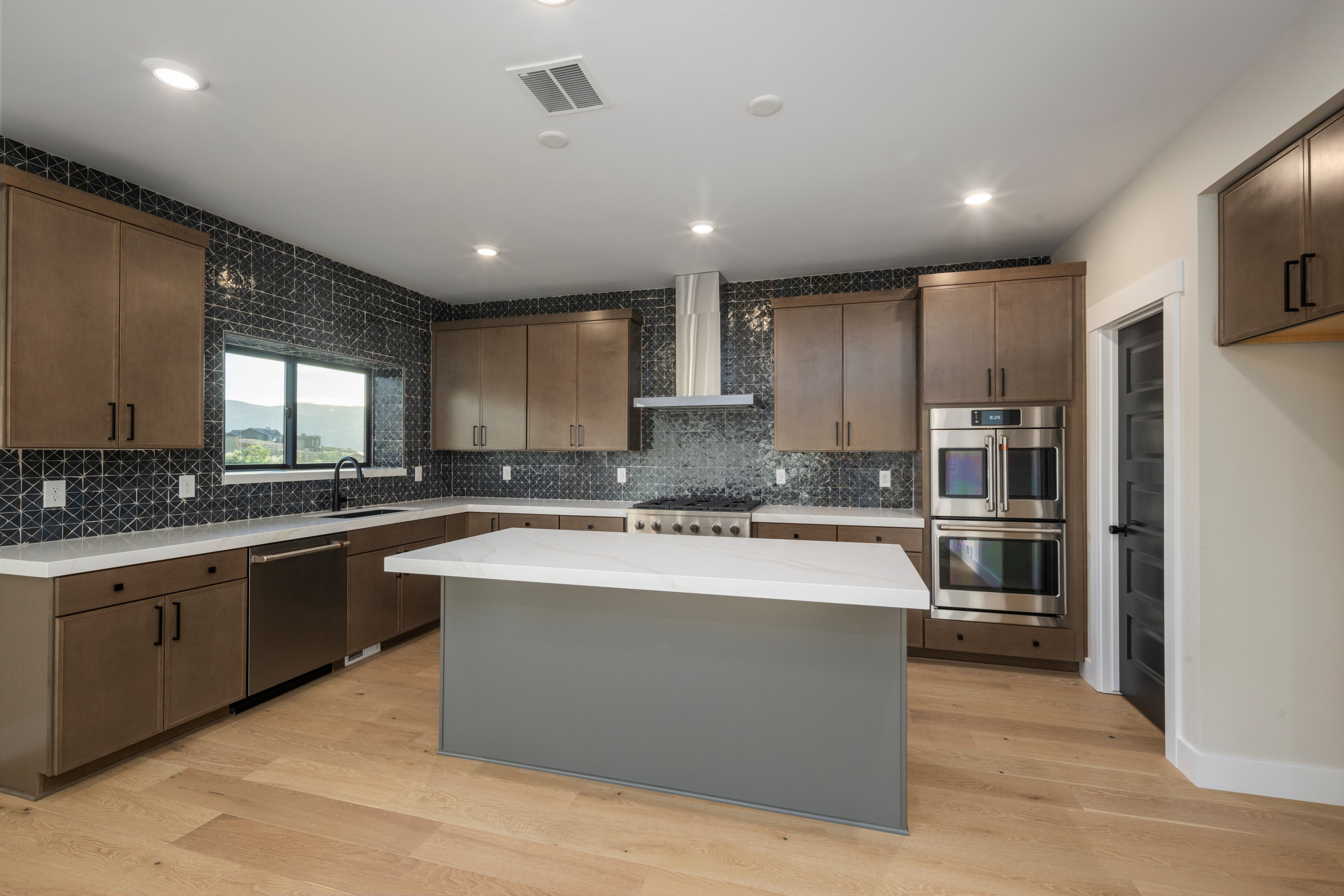 Modern kitchen featuring an island with a white solid surface countertop, light wood-finish flooring, brown cabinetry, stainless steel appliances, and a dark geometric tile backsplash
