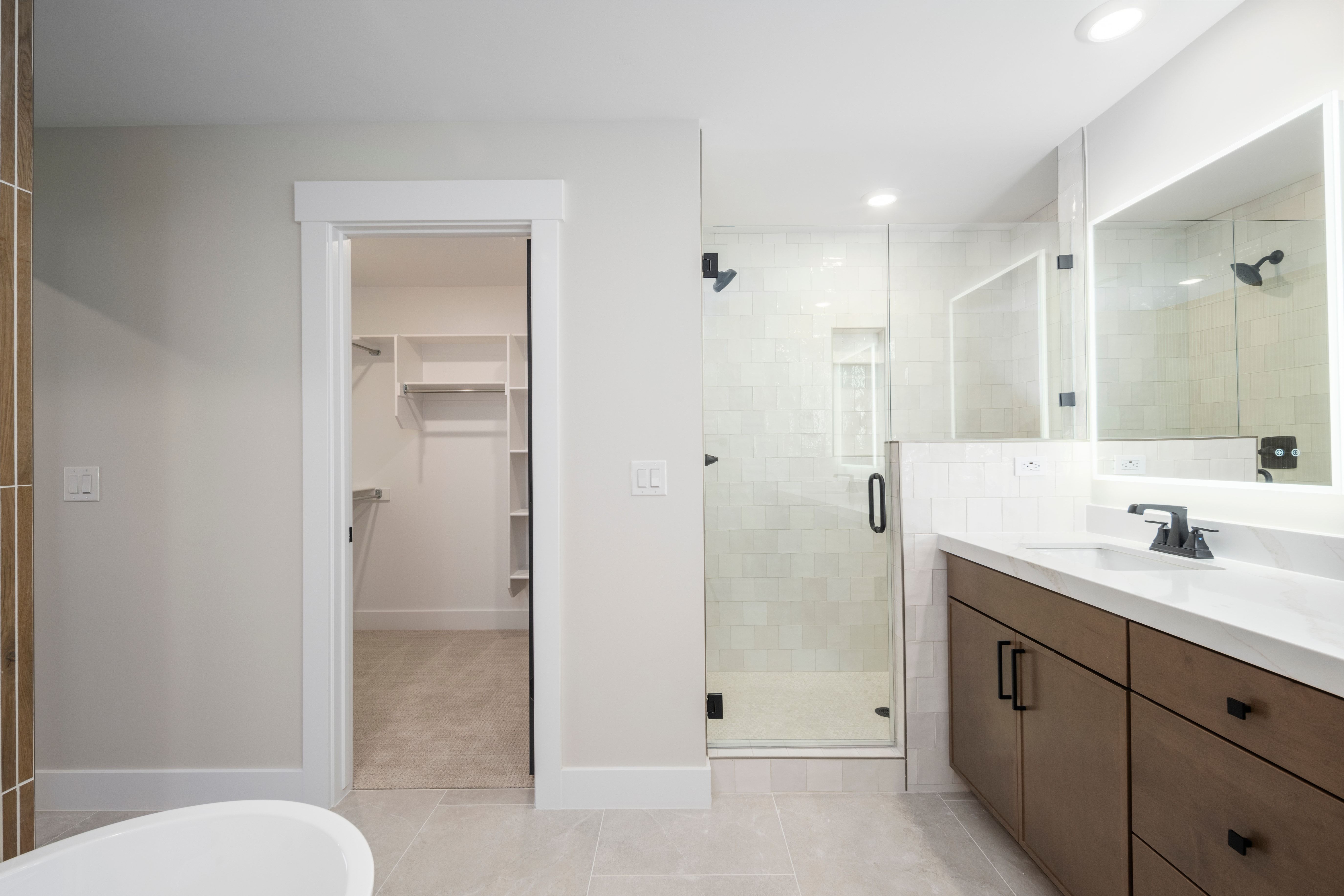 Bathroom featuring a walk-in shower with glass enclosure, single vanity with wood-finish cabinetry, illuminated mirror, and light-toned tile flooring