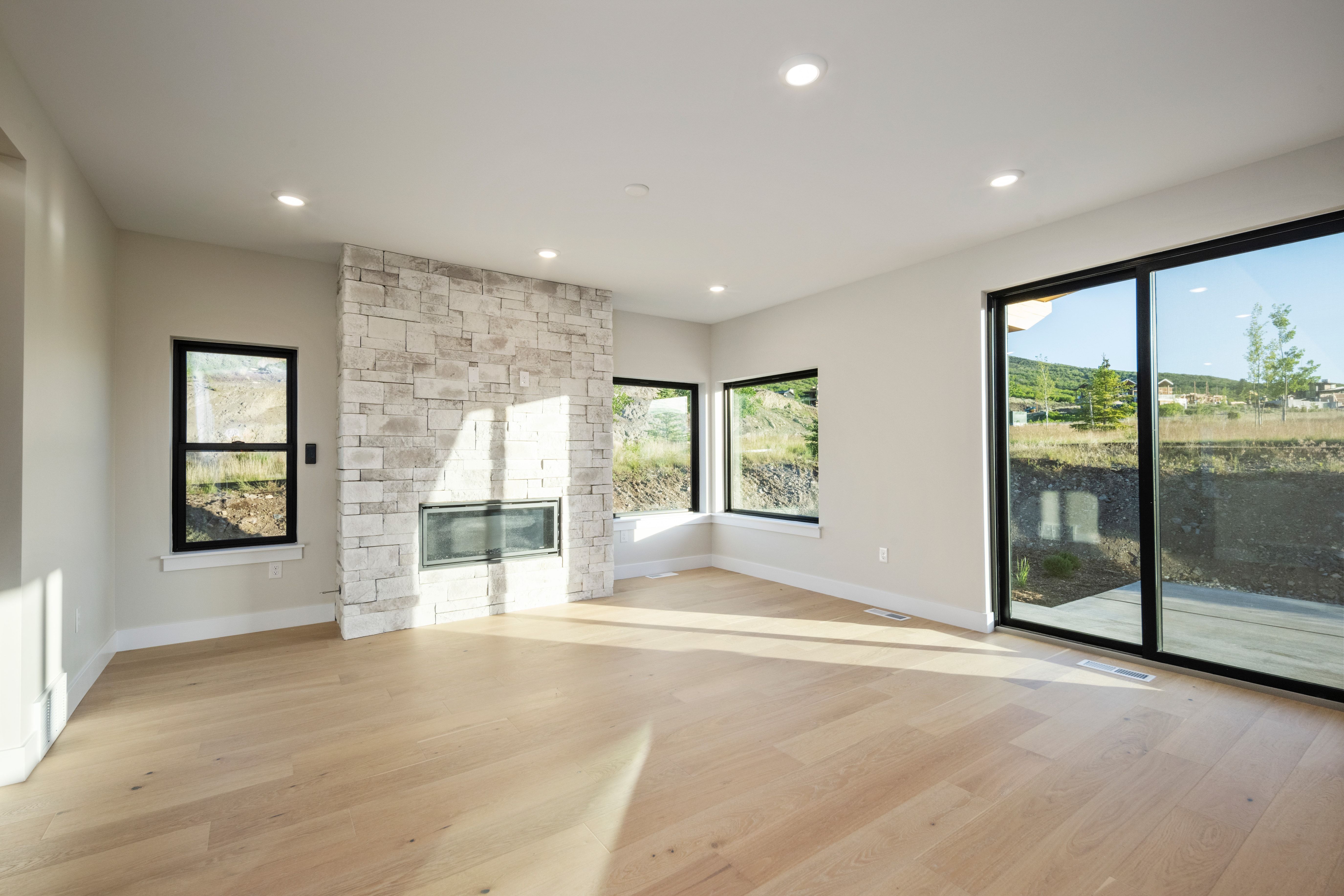 Spacious room featuring light wood-finish flooring, a stone-clad fireplace with a dark-framed insert, multiple large windows, a sliding glass door, and recessed ceiling lighting