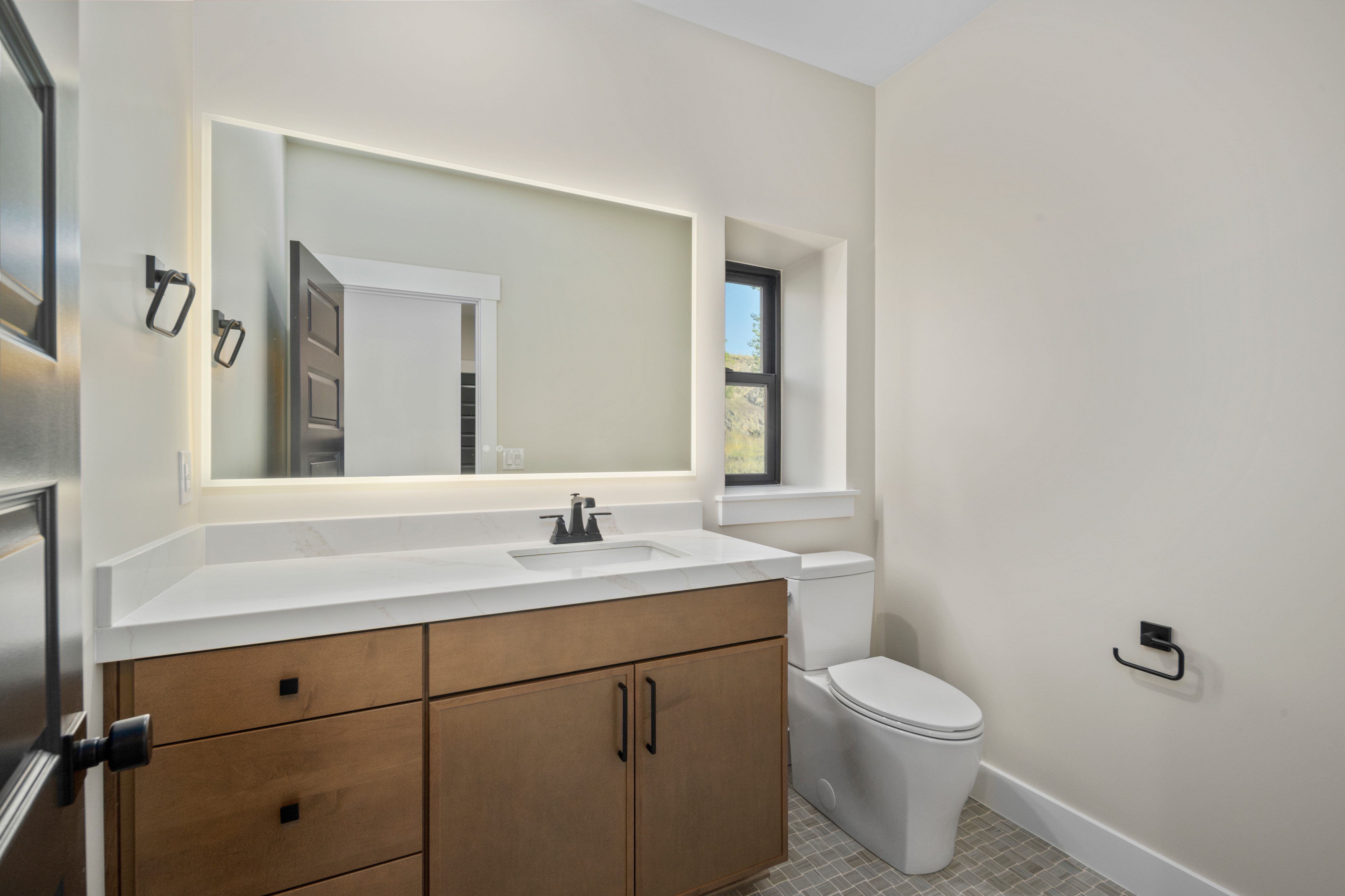 Bathroom featuring a wood-finish vanity with a white countertop, an integrated sink, a backlit mirror, and a window
