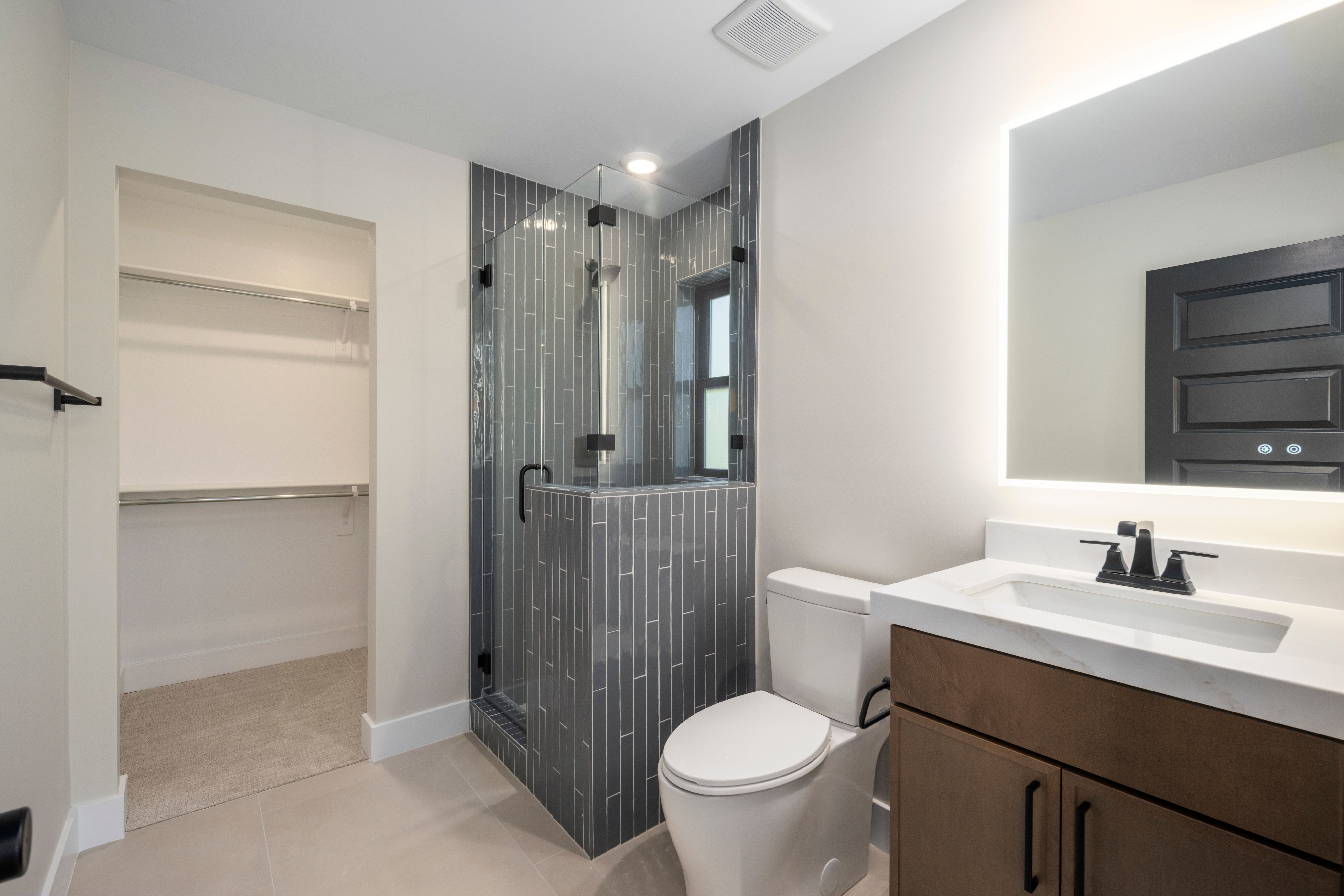 Bathroom featuring a glass-enclosed shower with vertical gray tile, a wood-finish vanity with a white countertop and integrated sink, a backlit mirror, and a walk-in closet with shelving and hanging rods