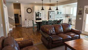 Open-concept living area featuring wood-finish flooring, a kitchen island with seating, white cabinetry, stainless steel appliances, and pendant lighting