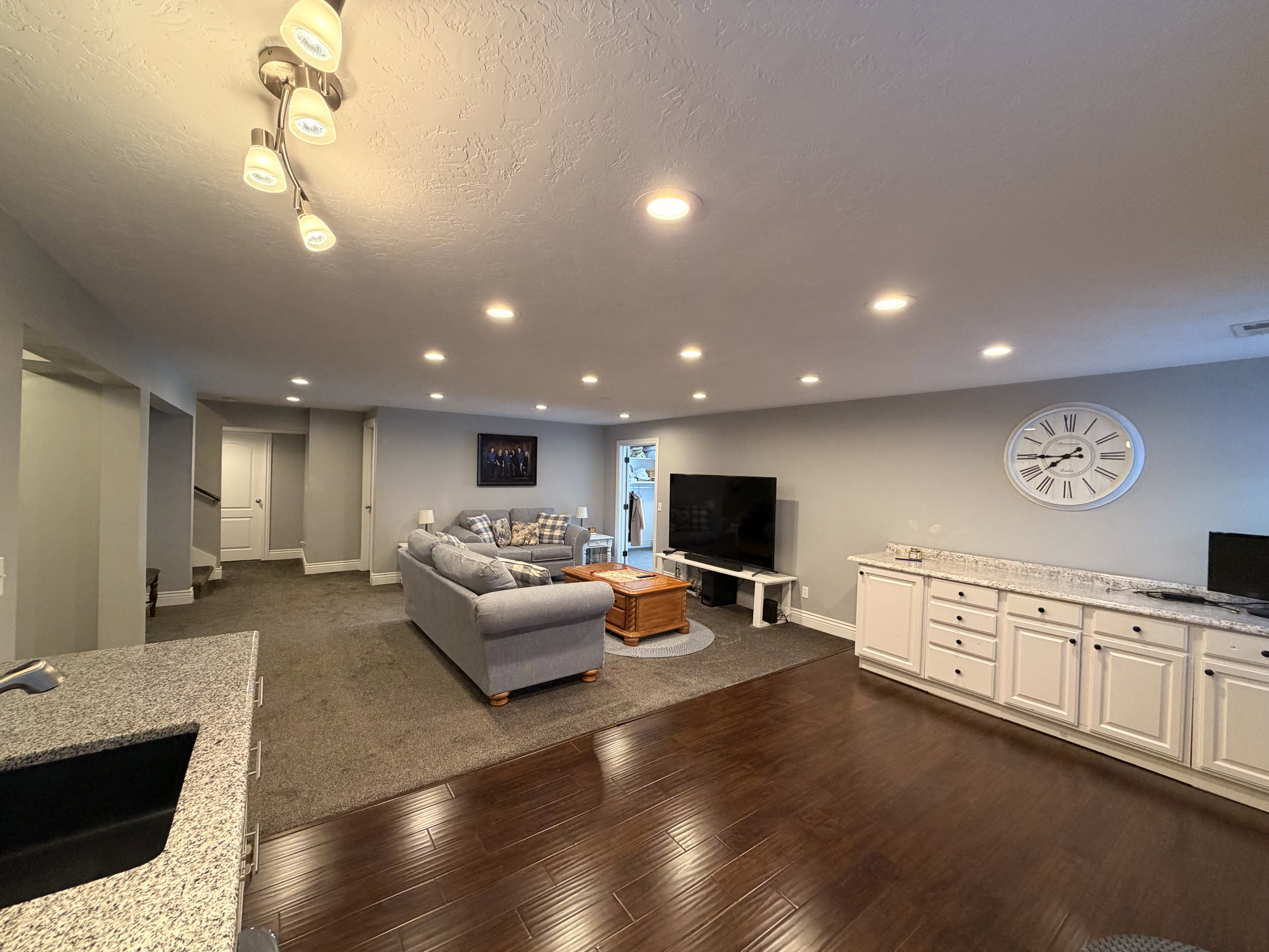 Open-concept living area featuring rich wood-finish flooring, recessed lighting, white cabinetry with a light speckled countertop, and a dark sink with a chrome faucet