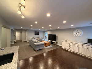 Open-concept living area featuring rich wood-finish flooring, recessed lighting, white cabinetry with a light speckled countertop, and a dark sink with a chrome faucet