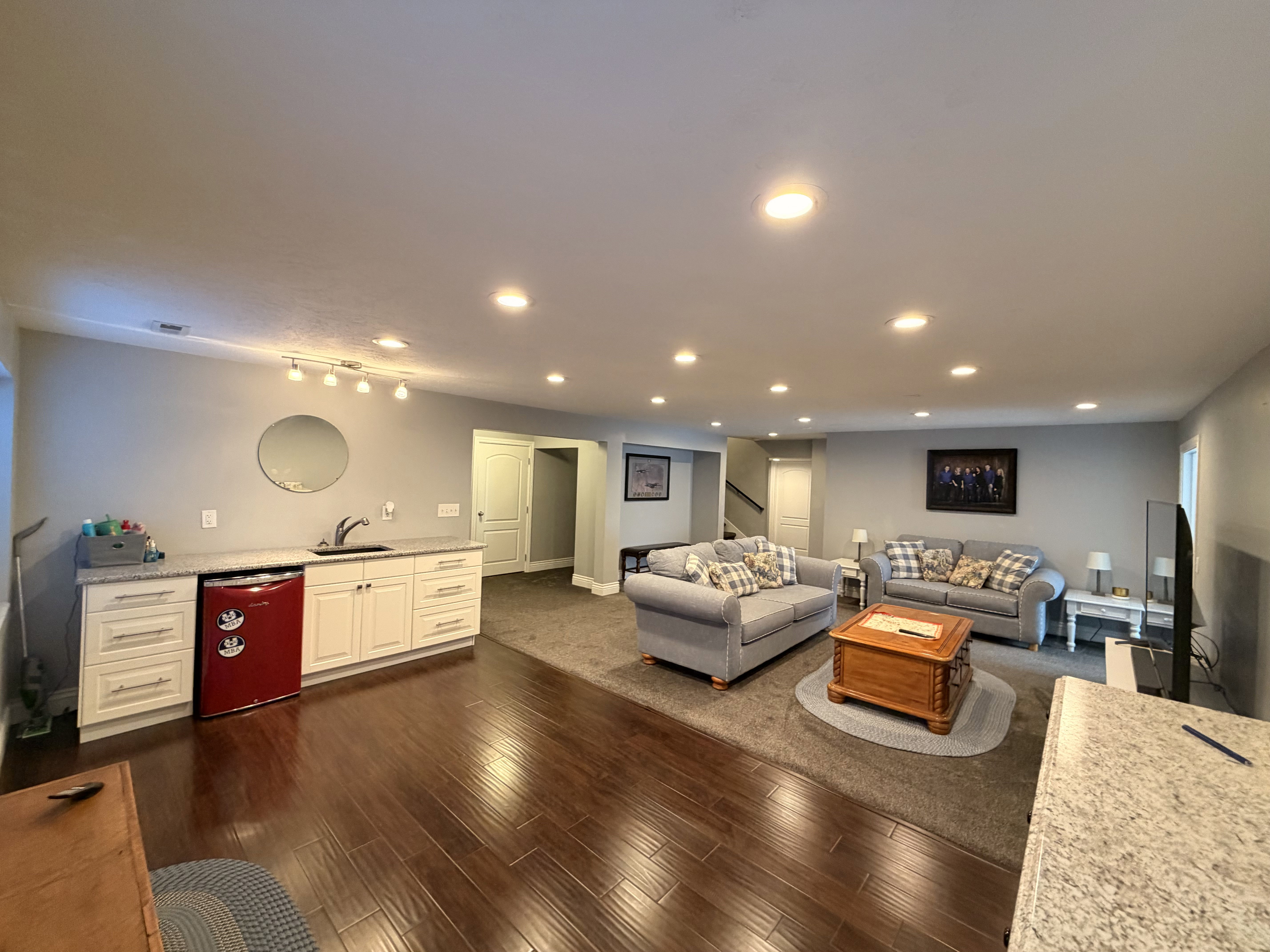 Spacious lower-level living area featuring recessed lighting, a wet bar with white cabinetry and stone-finish countertops, wood-finish flooring, and soft gray wall tones