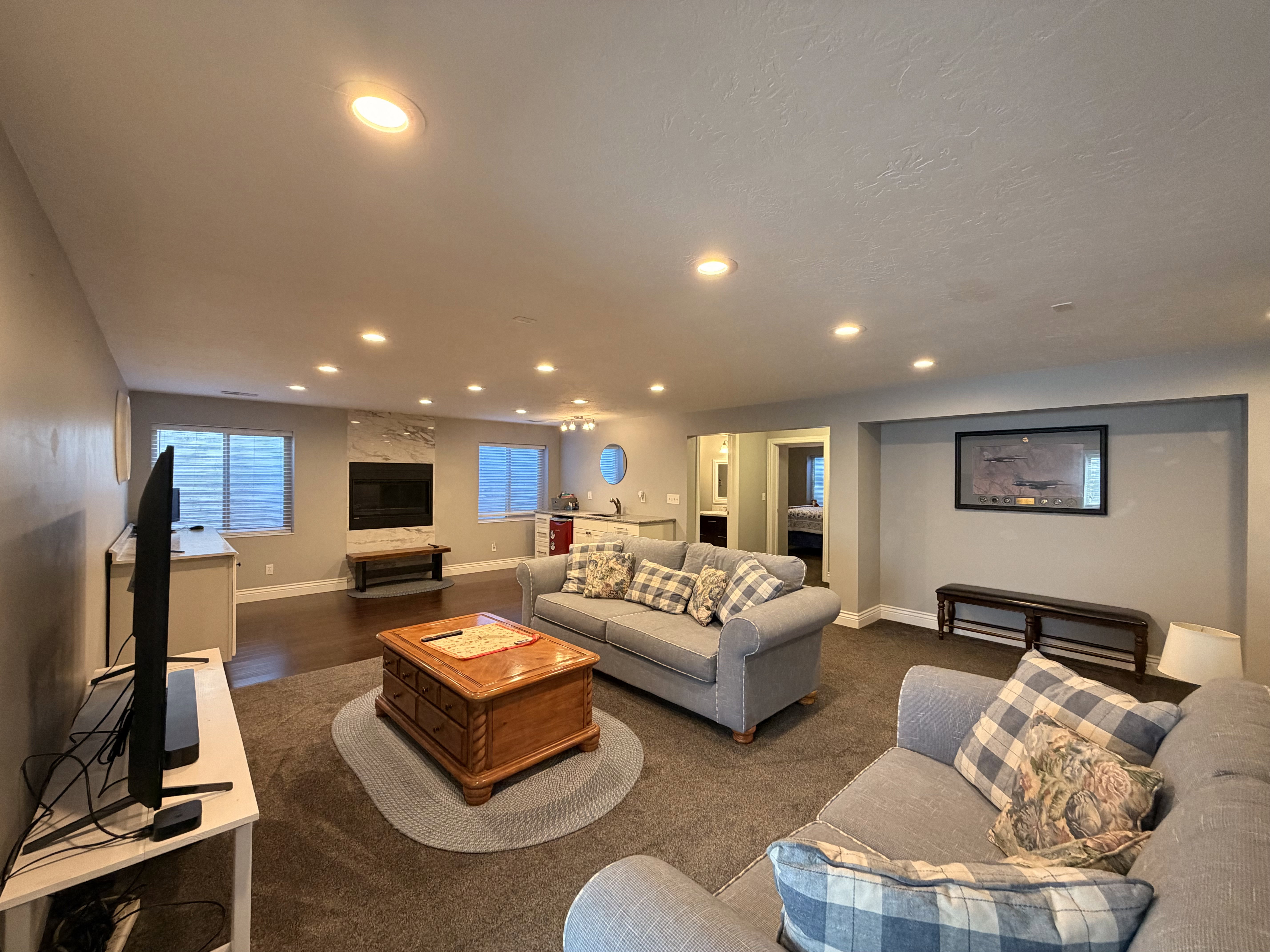Expansive living area featuring recessed lighting, a stone-clad fireplace with a dark wood mantel, and wood-finish flooring extending to a built-in wet bar