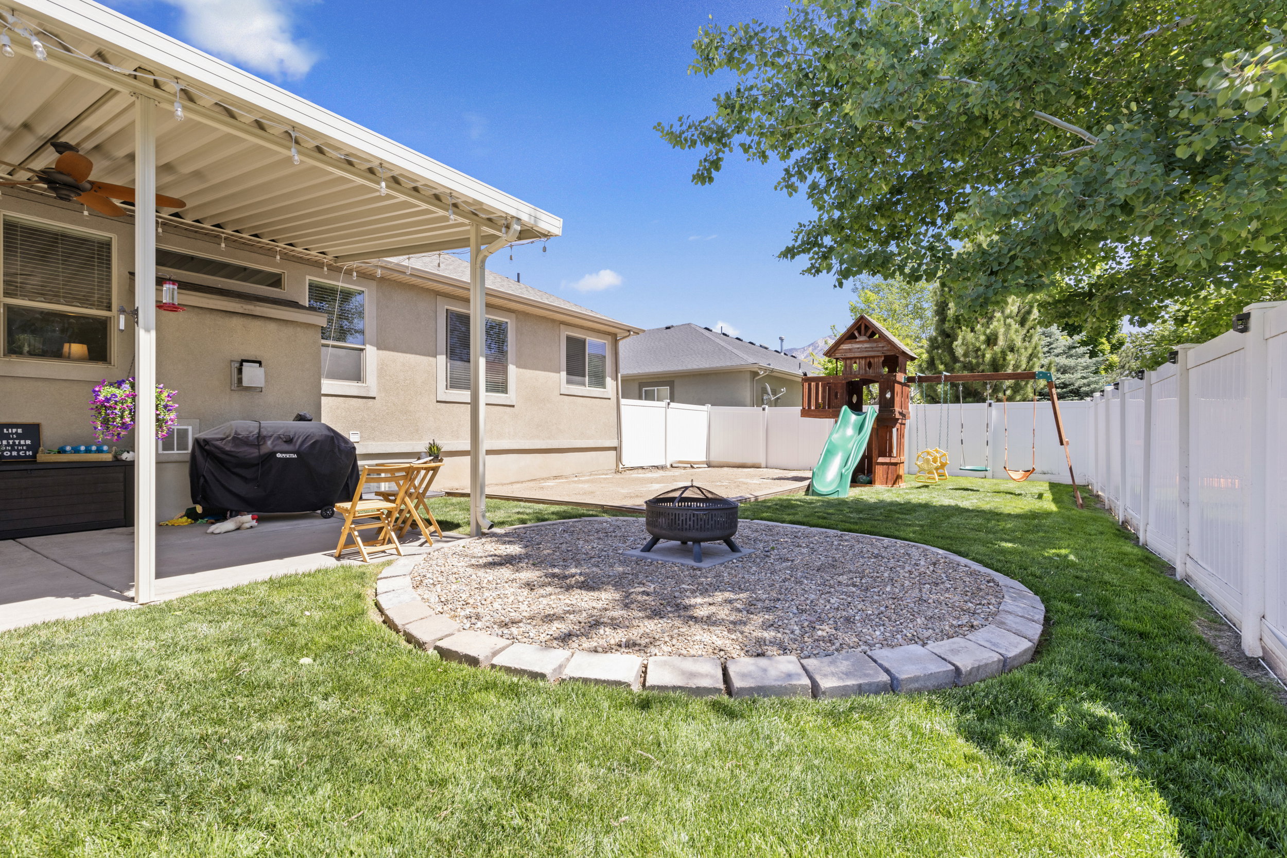 Expansive backyard featuring a covered patio, a defined gravel fire pit area with stone edging, established lawn, and white privacy fencing
