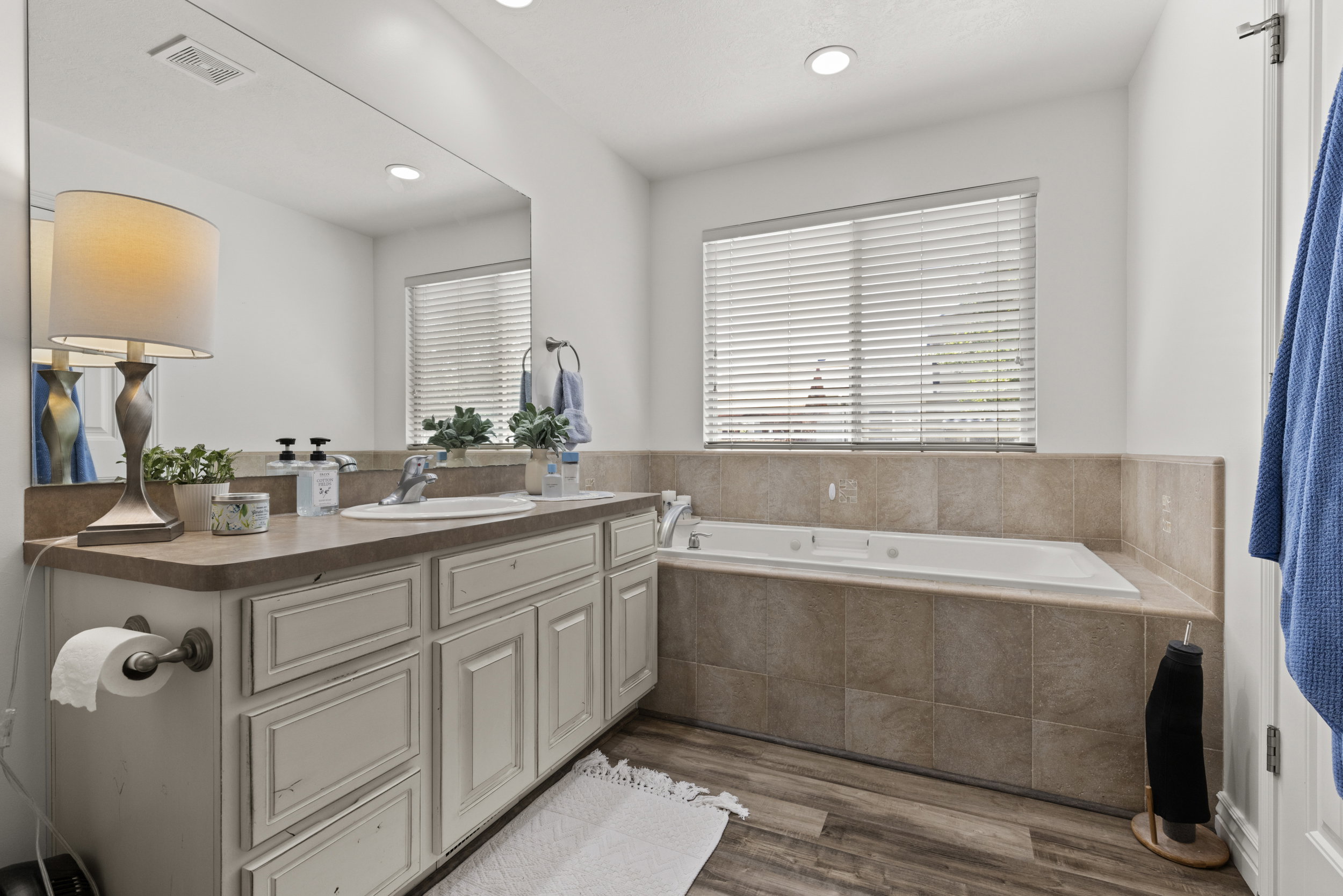 Bathroom featuring a white drop-in tub with surrounding tile, a window with blinds, a vanity with an integrated sink, a large wall mirror, and wood-finish flooring