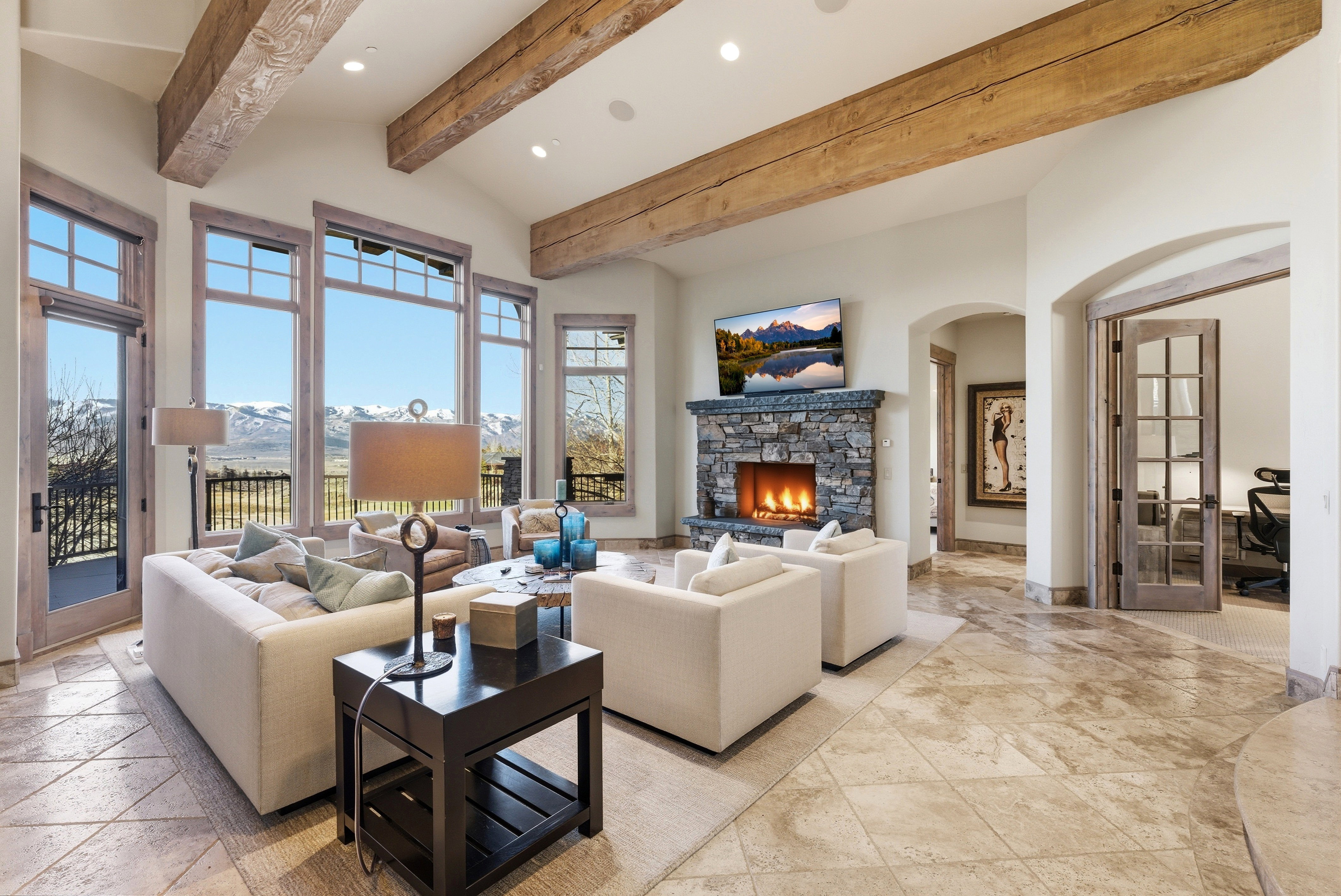 Great room featuring vaulted ceilings with exposed wood beams, a stone-clad fireplace, large windows, and tile flooring