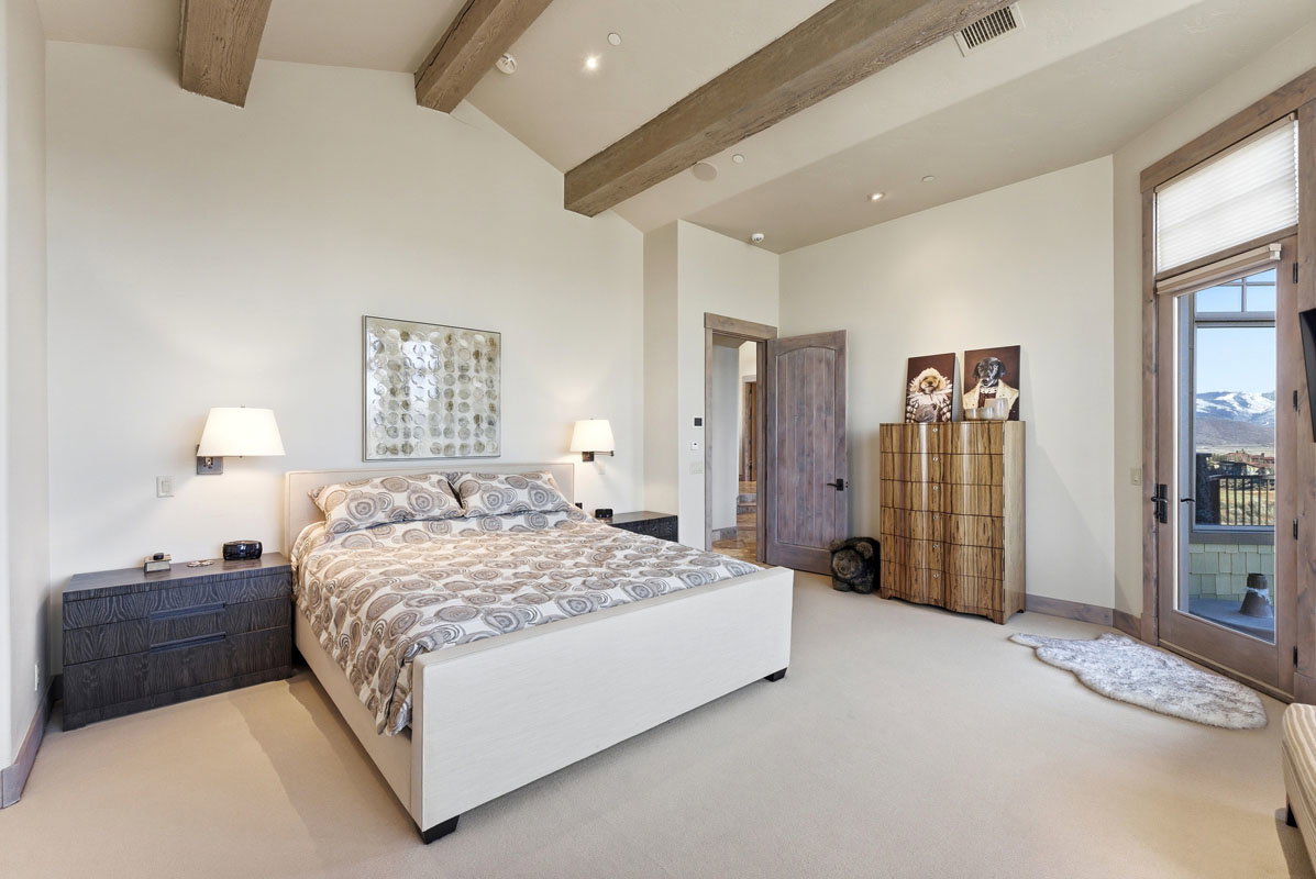 Vaulted ceiling with exposed wood beams, wall-mounted sconces, recessed lighting, wood-finish flooring, and a glass-paneled door leading to an outdoor space with mountain scenery