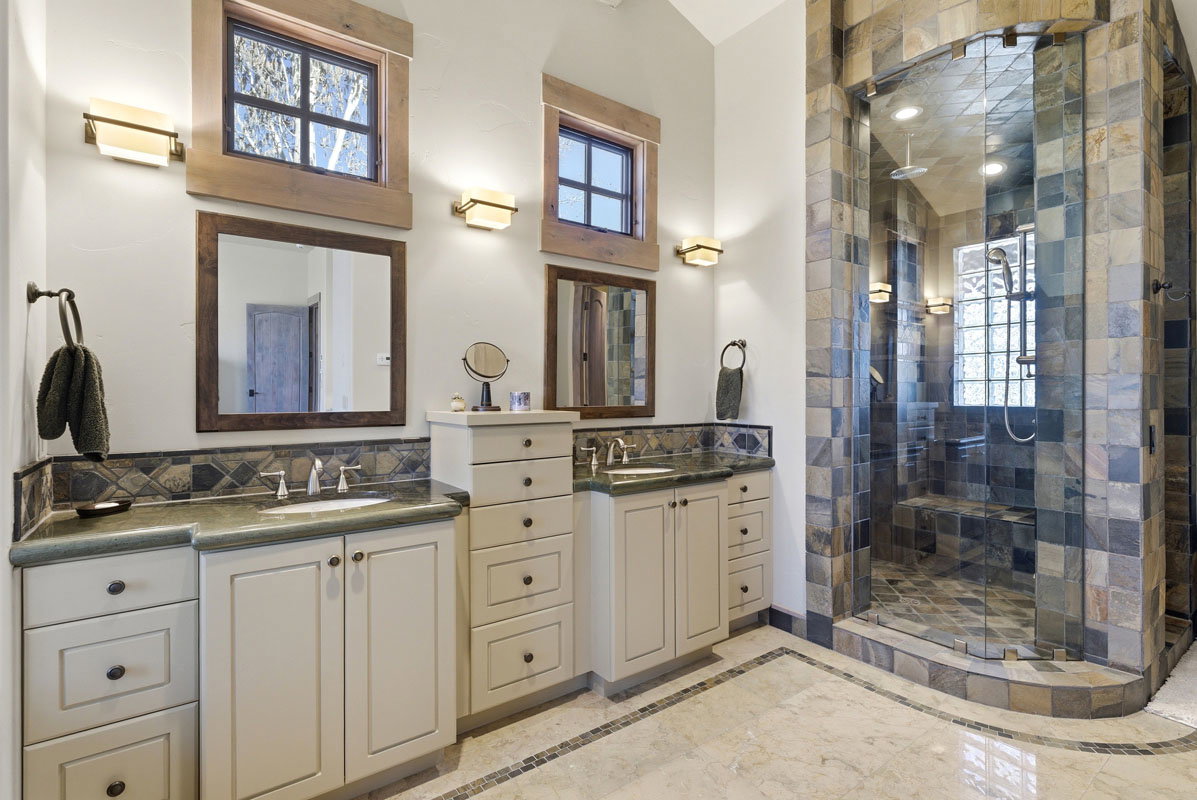 Spacious bathroom featuring a glass-enclosed shower with stone tile surround, dual vanity areas with undermount sinks, and light-toned cabinetry