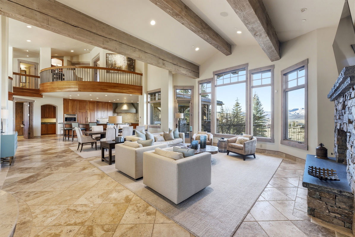 Spacious living area featuring exposed wood beams, a stone fireplace, large picture windows, and patterned tile flooring