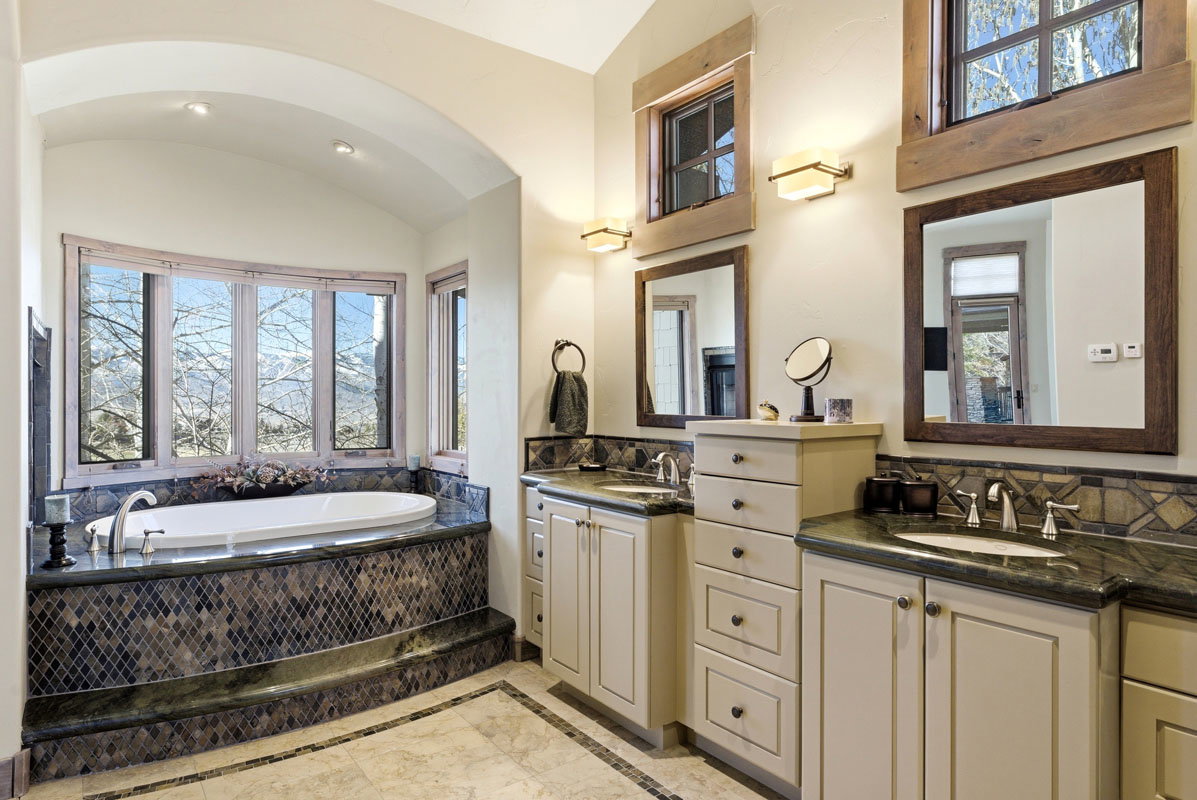 Soaking tub with a tiled surround, dual vanities with stone countertops, and multiple windows