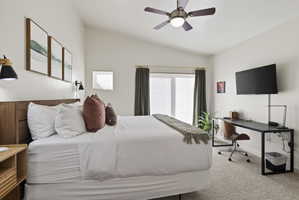 Vaulted ceiling bedroom featuring a wood-finish headboard, a ceiling fan with light fixture, wall-mounted sconces, and a large window with privacy blinds