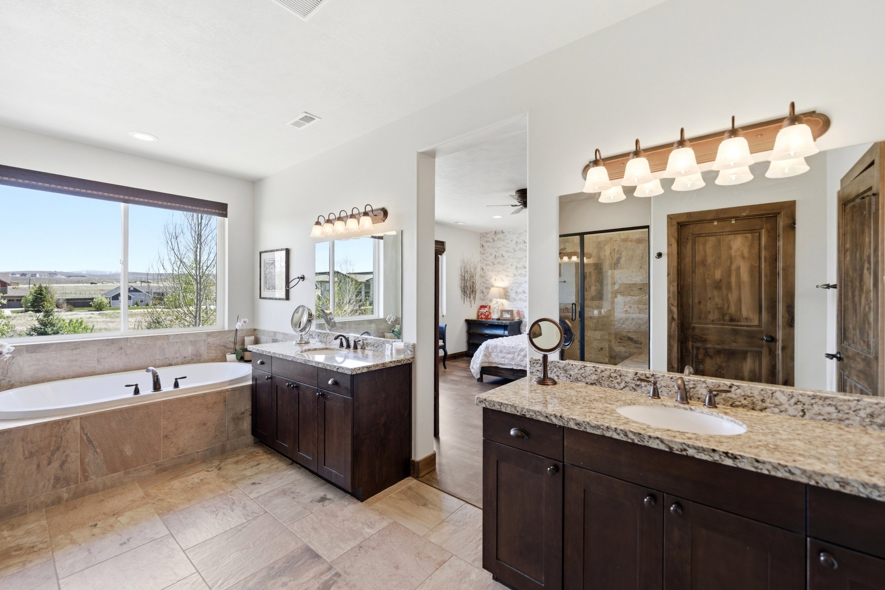 Spacious bathroom featuring a built-in soaking tub with a large picture window, stone-look tile flooring, and dark wood vanities with light granite countertops