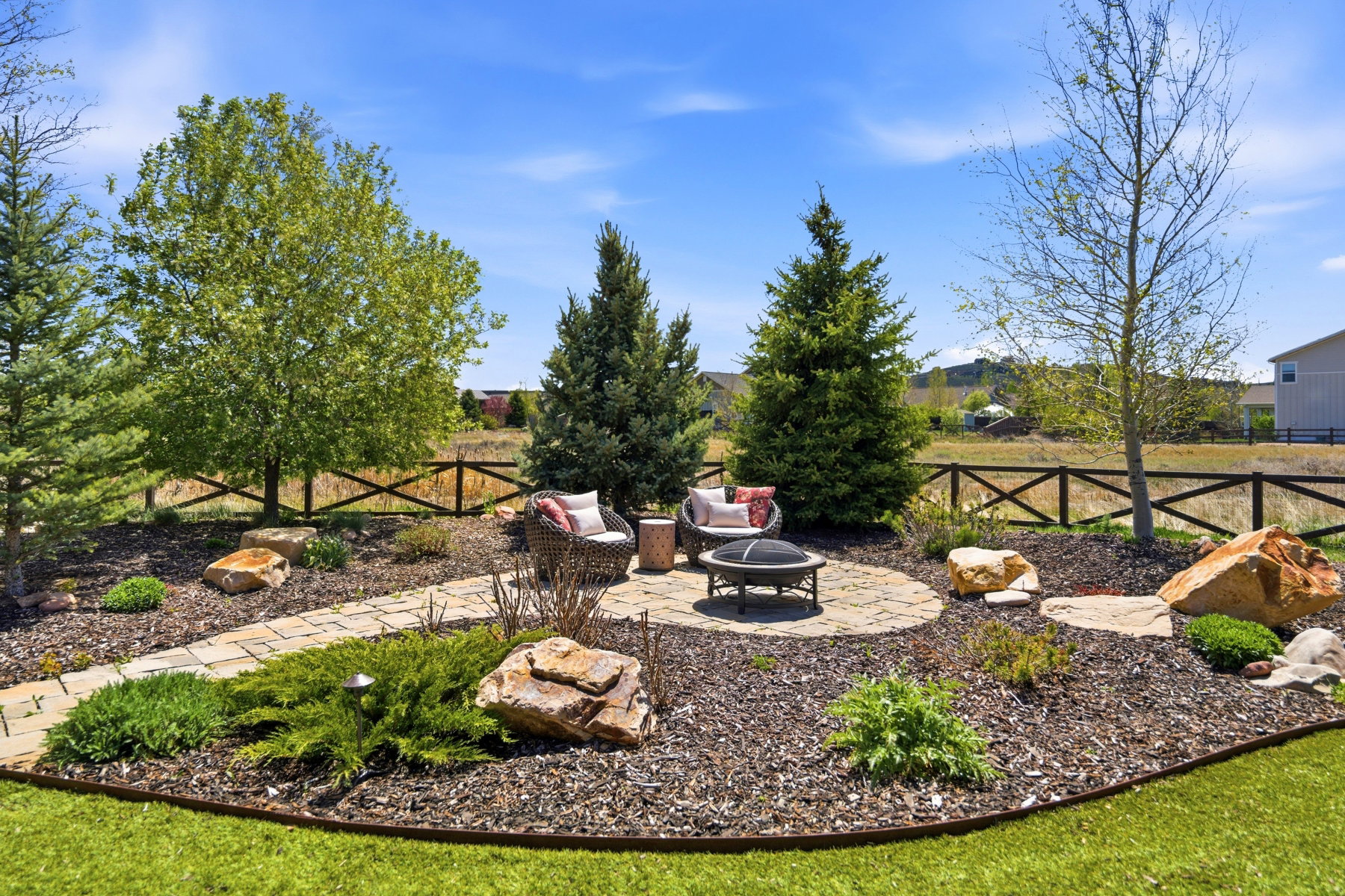 Landscaped outdoor area featuring a circular stone patio with a fire pit, bordered by natural rock formations and a variety of mature evergreen and deciduous trees