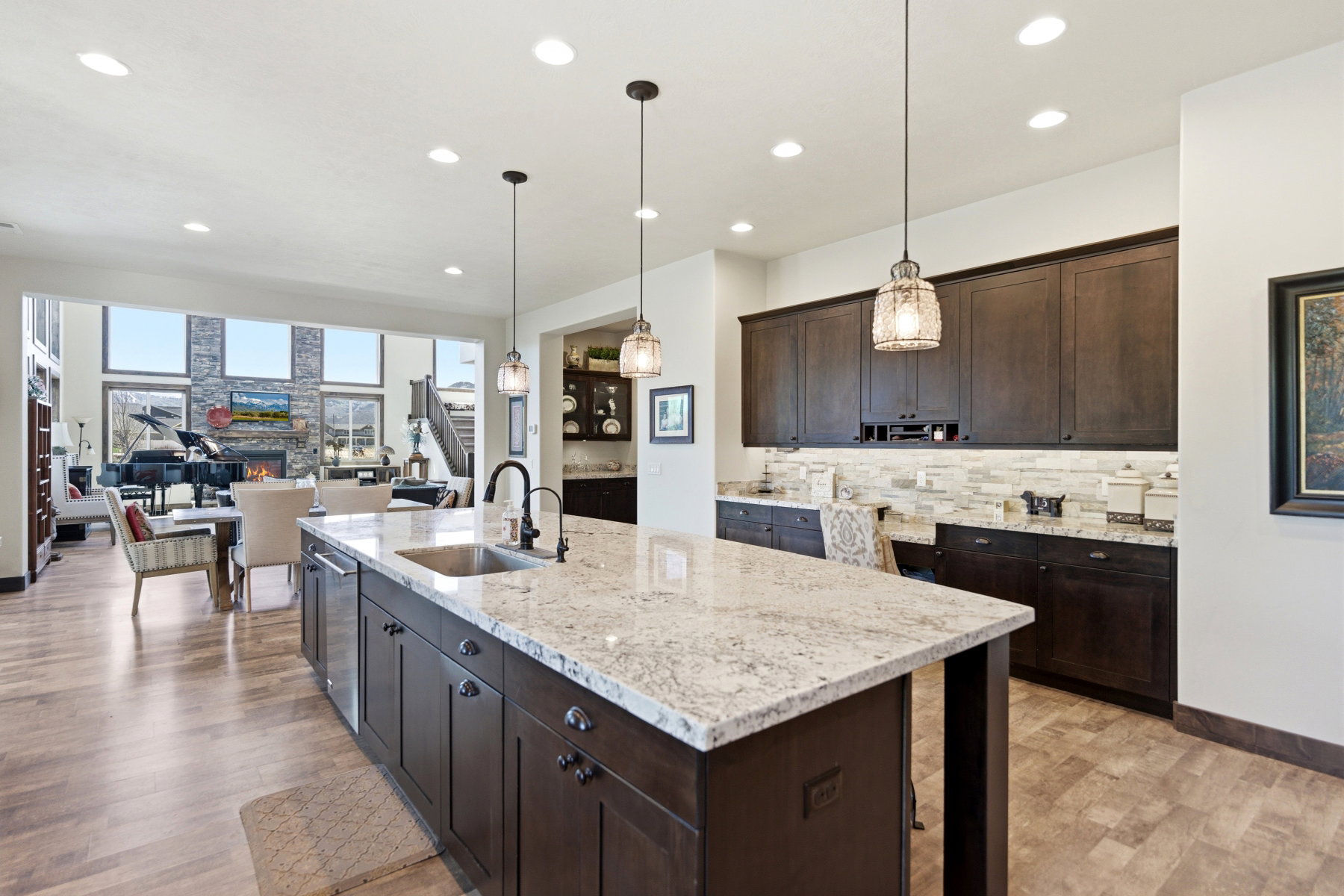 Spacious kitchen featuring a large island with a stainless steel sink and a dark wood-finish base, complemented by dark wood-finish cabinetry, light-toned countertops, and a stone-tiled backsplash