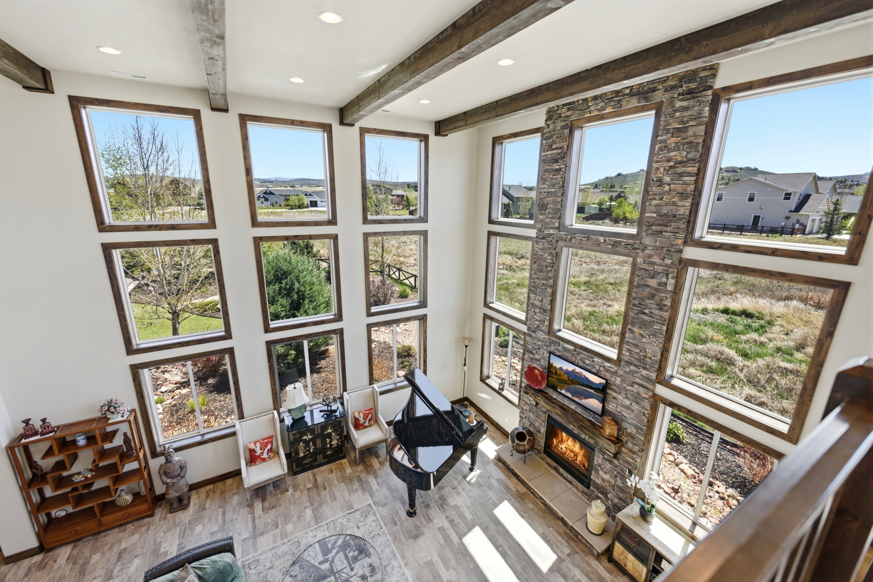 Two-story living area featuring a stone fireplace, wood-finish flooring, exposed ceiling beams, and multiple picture windows with wood trim