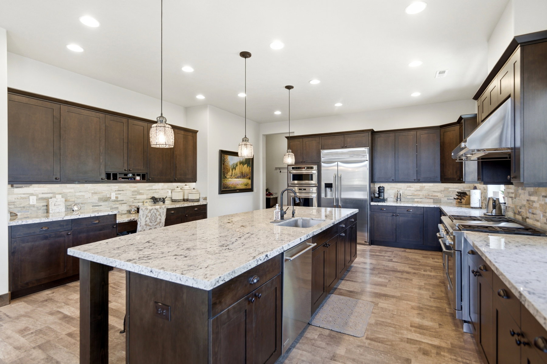 Expansive kitchen featuring a large central island with a sink, stainless steel appliances, dark wood-finish cabinetry, light-toned countertops, and a stone tile backsplash