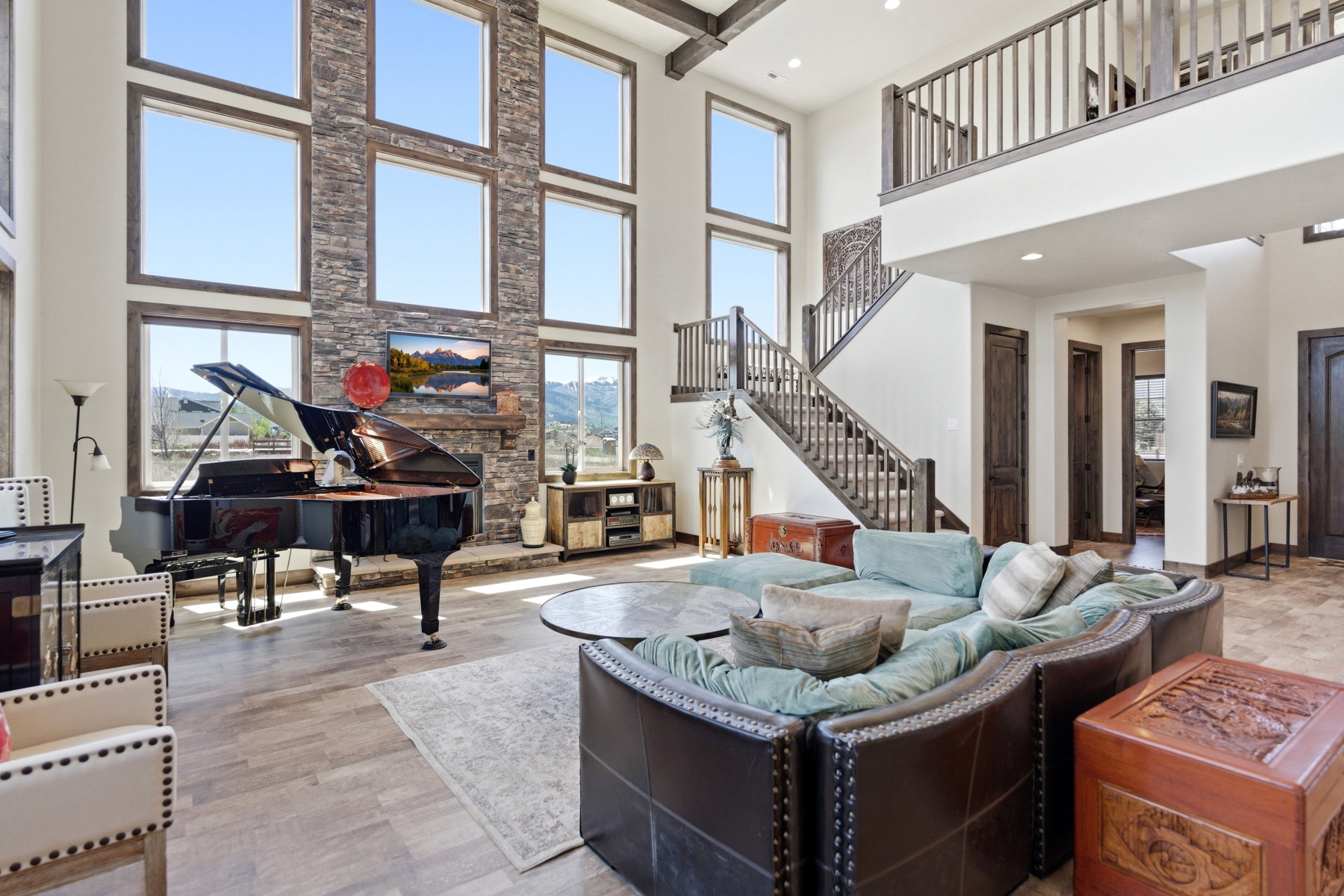 Soaring two-story living area featuring a stacked stone fireplace with a wood mantle, an array of tall windows, wood-finish flooring, and a multi-level staircase with wood railing