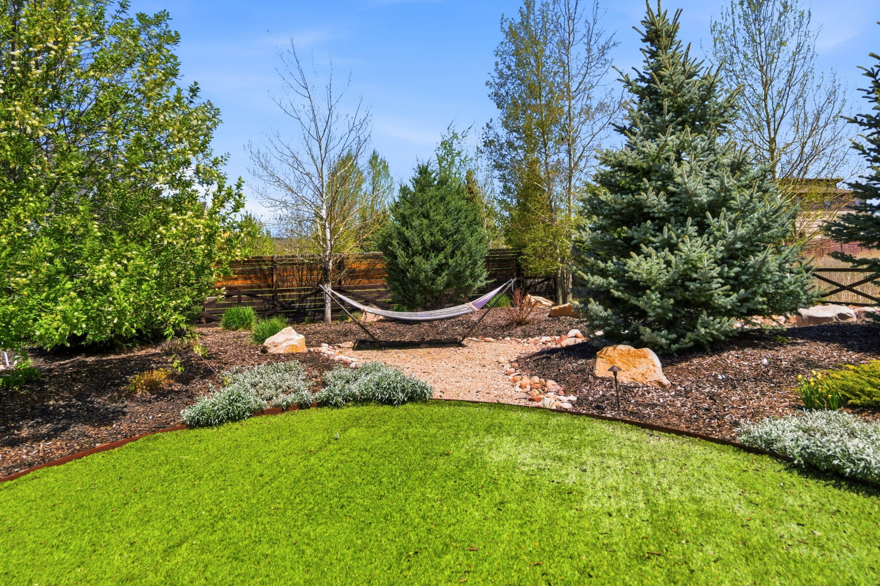Landscaped backyard featuring an artificial turf lawn, a gravel pathway, mature trees, and natural rock accents
