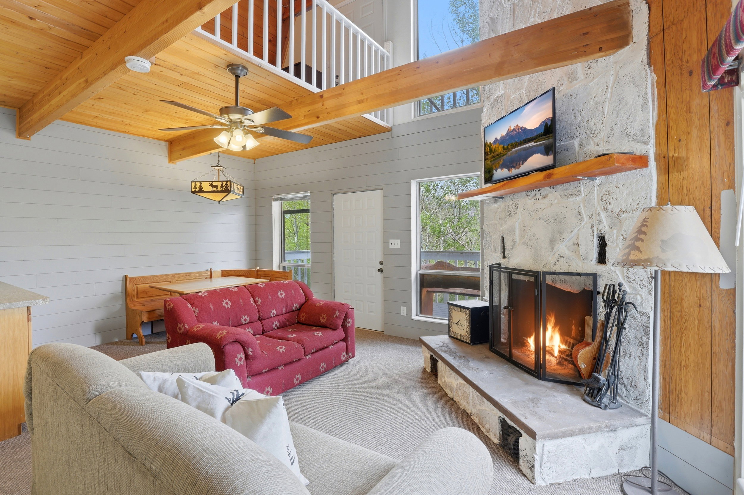 Living area featuring a stone fireplace with a raised hearth, wood-finish flooring, exposed wood ceiling beams, and a mezzanine with white railings