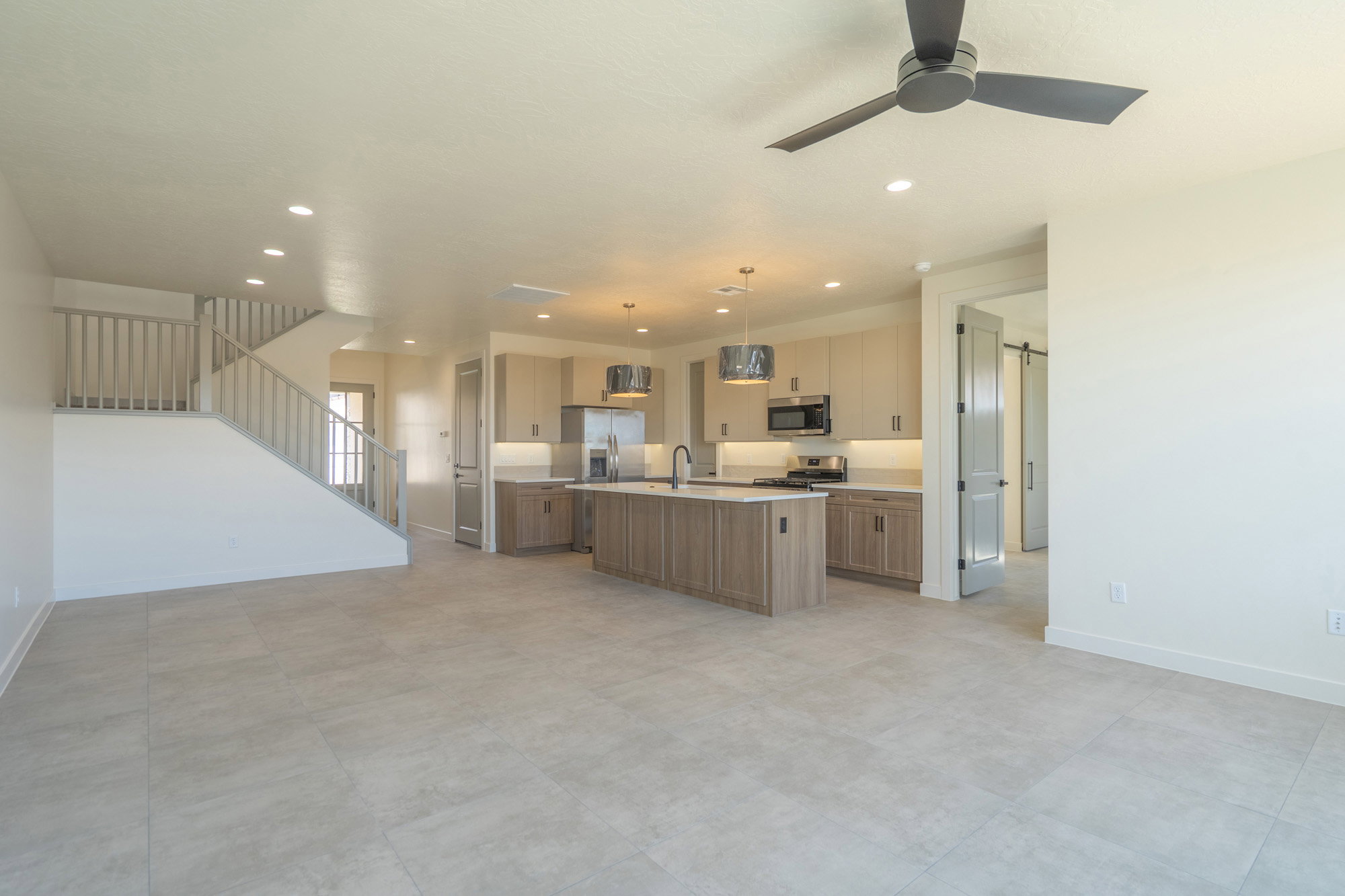 Open-concept living area featuring light-toned tile flooring, a contemporary kitchen with wood-finish cabinetry, a central island with an undermount sink, stainless steel appliances, and a modern ceiling fan