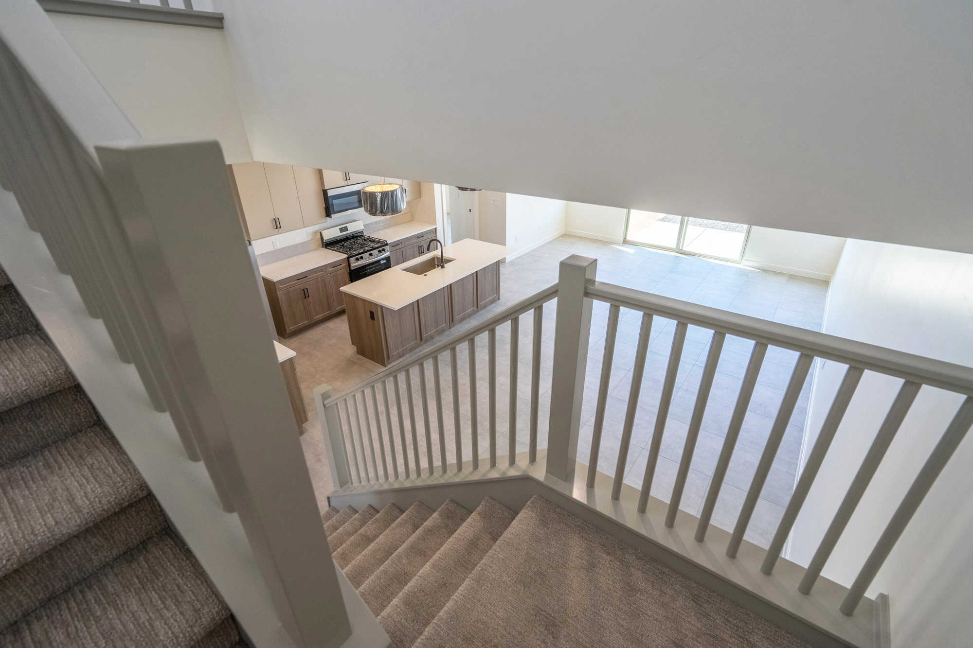 Carpeted staircase with white railings overlooking a modern kitchen featuring wood-finish cabinetry, light countertops, stainless steel appliances, and a central island with a sink