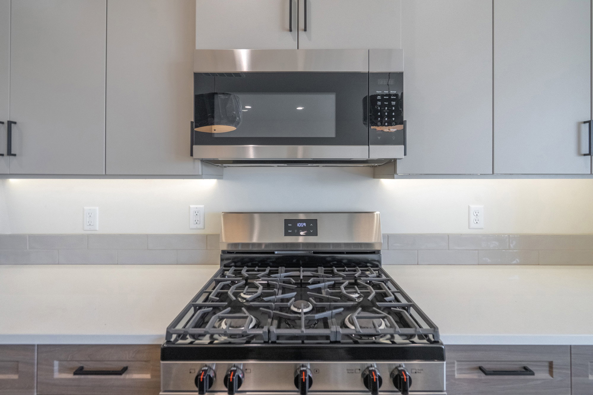 Stainless steel gas range with an overhead microwave, gray flat-panel cabinetry, under-cabinet lighting, and light-toned countertops