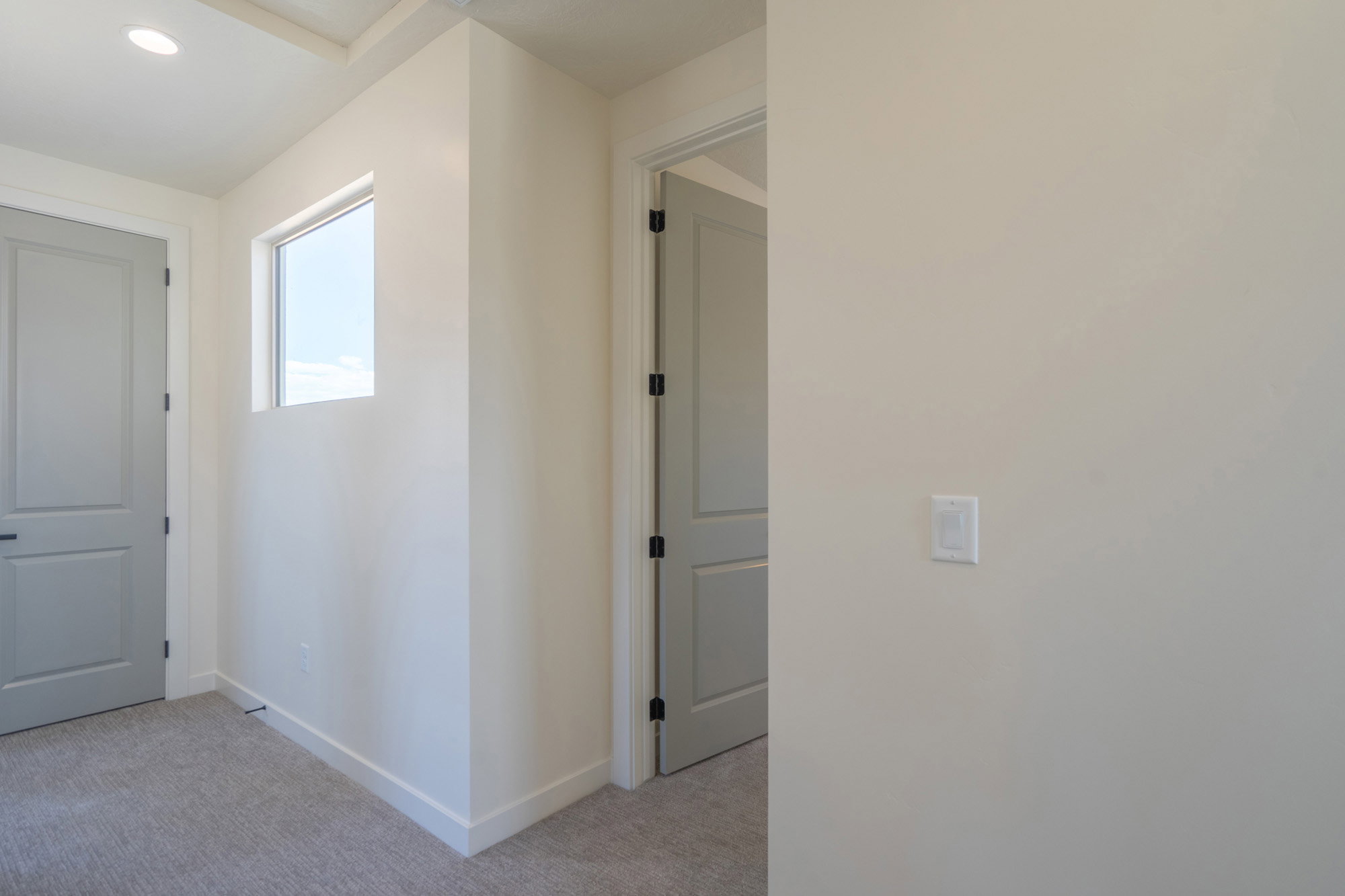 Interior hallway featuring light neutral wall paint, gray paneled doors with black hardware, a high-set window, recessed ceiling lighting, and light-colored carpeting