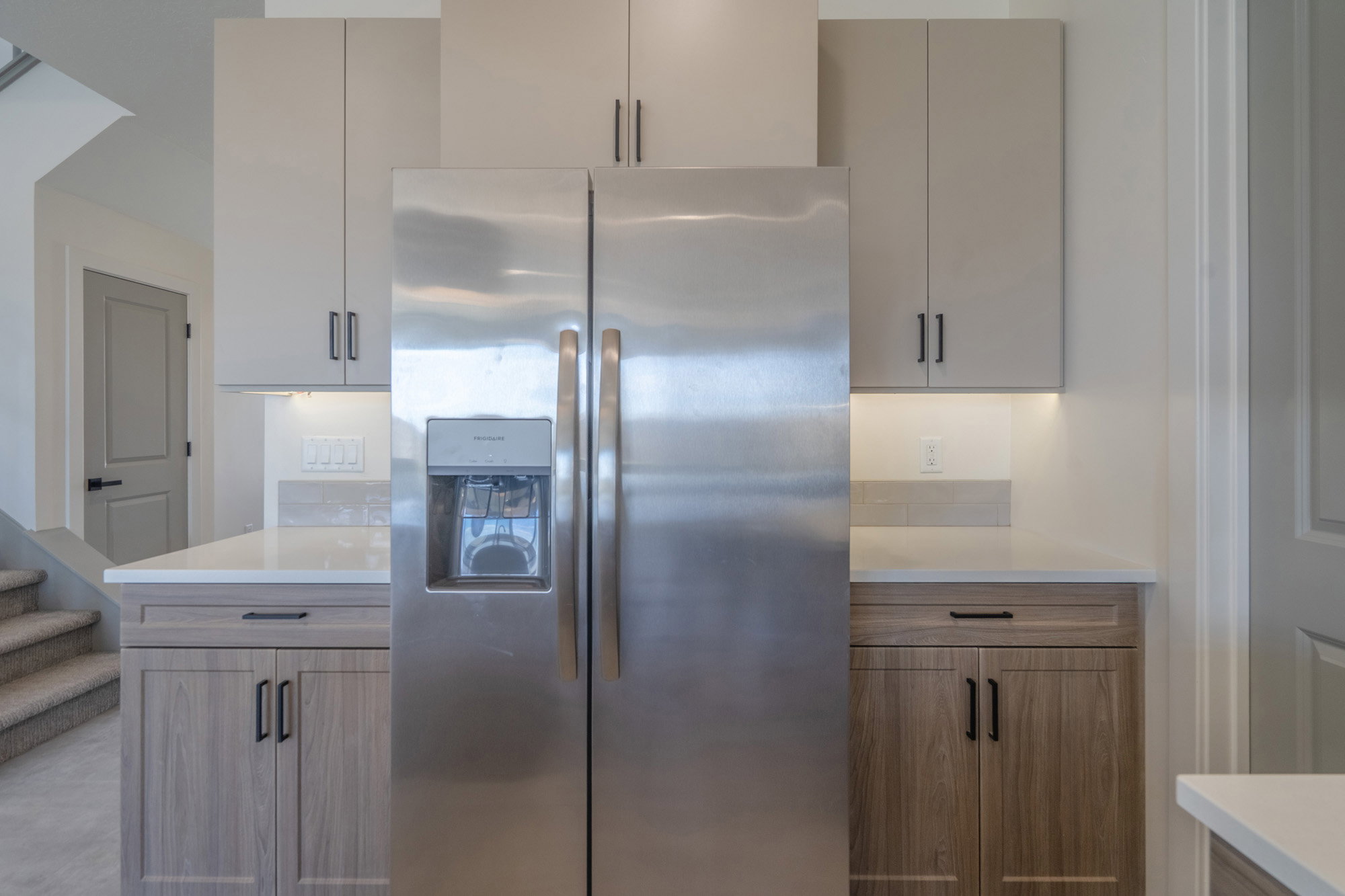 Stainless steel refrigerator with a dispenser, integrated into a kitchen featuring light wood-finish cabinetry, white countertops, and a subway tile backsplash