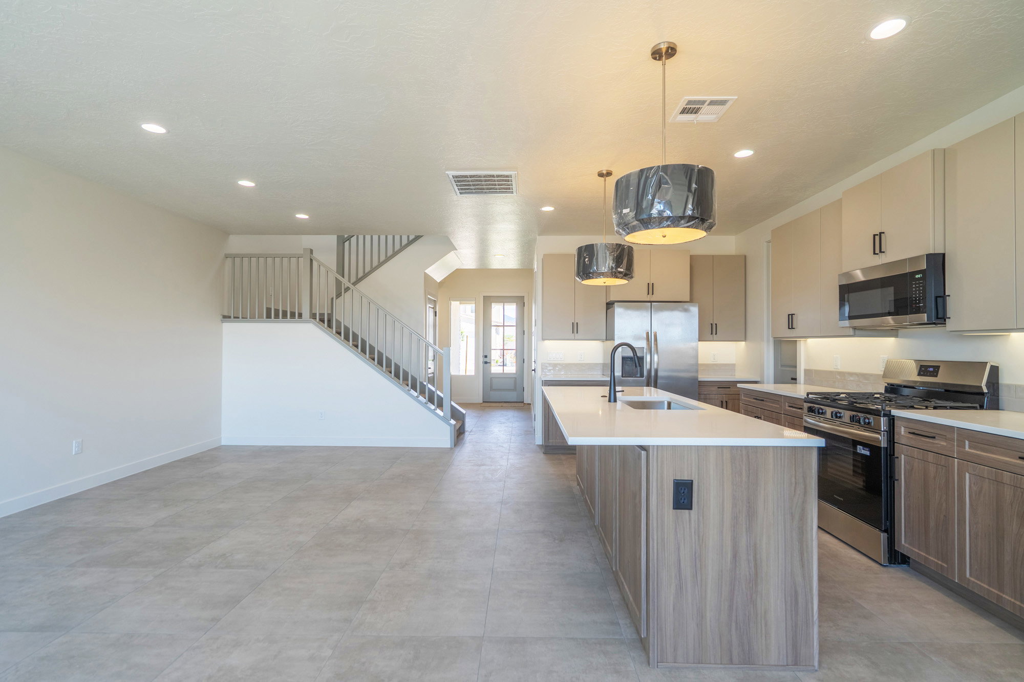 Open-concept living space with light-tone tile flooring, featuring a modern kitchen island with integrated sink, stainless steel appliances, and a staircase with light wood banisters