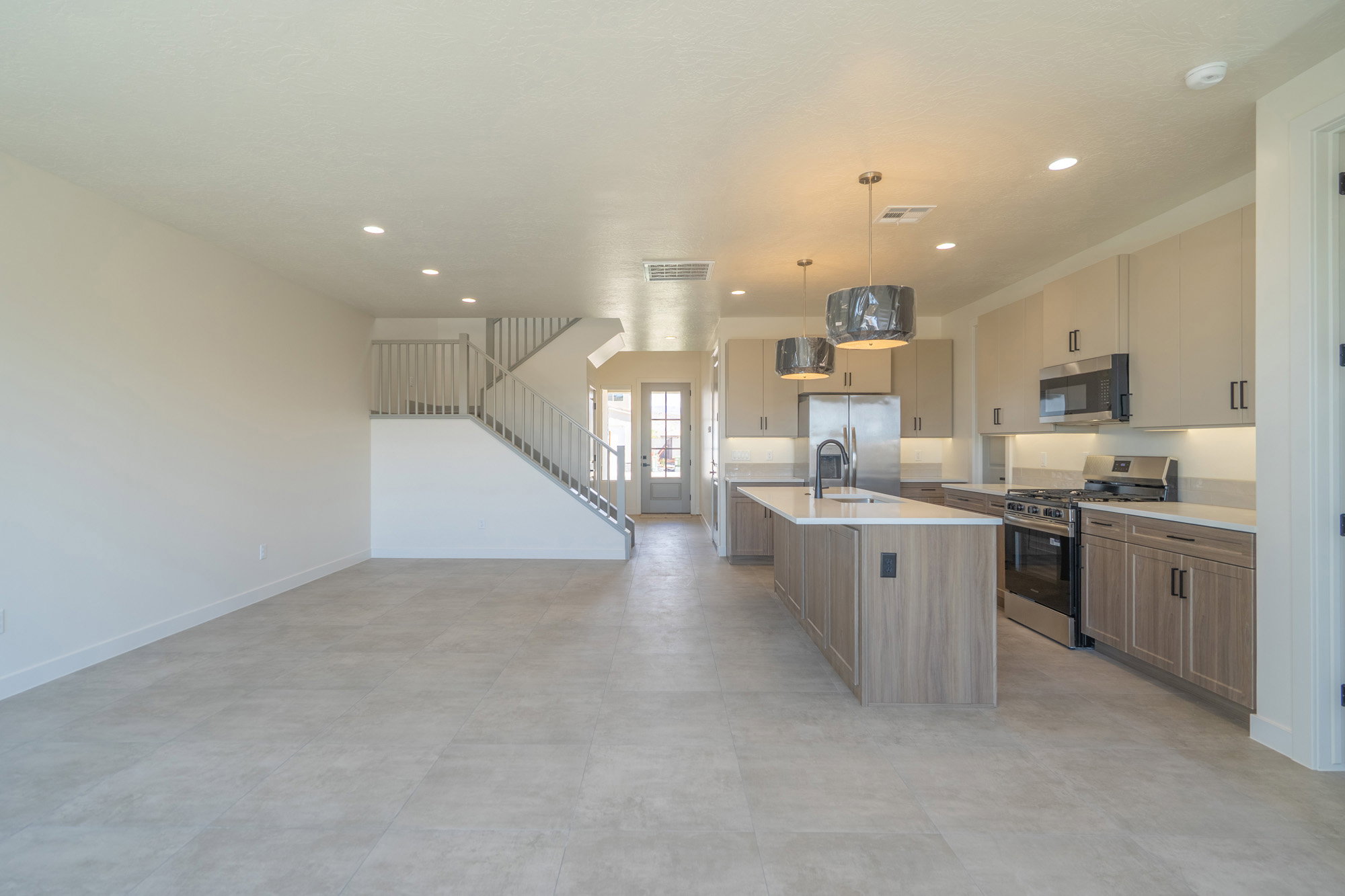 Open-concept living area featuring light-toned tile flooring, a modern kitchen with a center island, stainless steel appliances, and two-tone cabinetry, alongside a contemporary staircase with light wood banisters