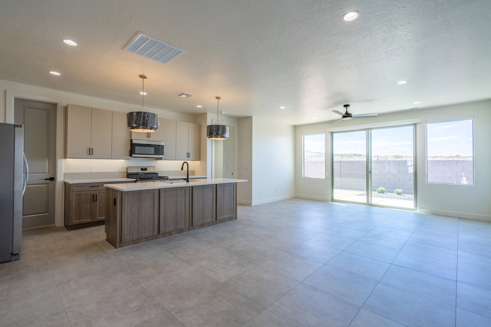 Open-concept living space featuring light tile flooring throughout, a modern kitchen island with an integrated sink, stainless steel appliances, and ample natural light from extensive sliding glass doors and windows
