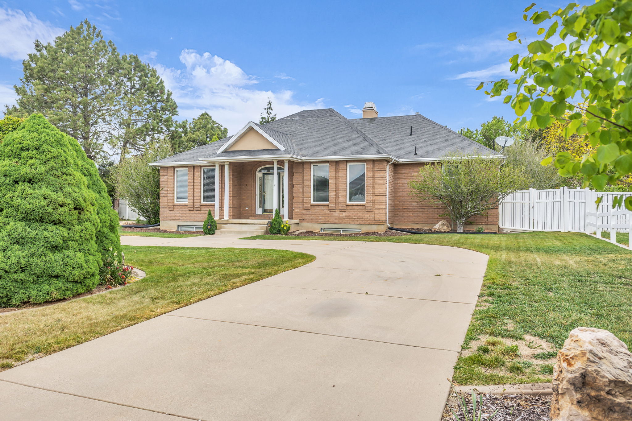 Brick exterior with a prominent portico entry, large front windows, a dark shingle roof, a concrete driveway, and a white fence