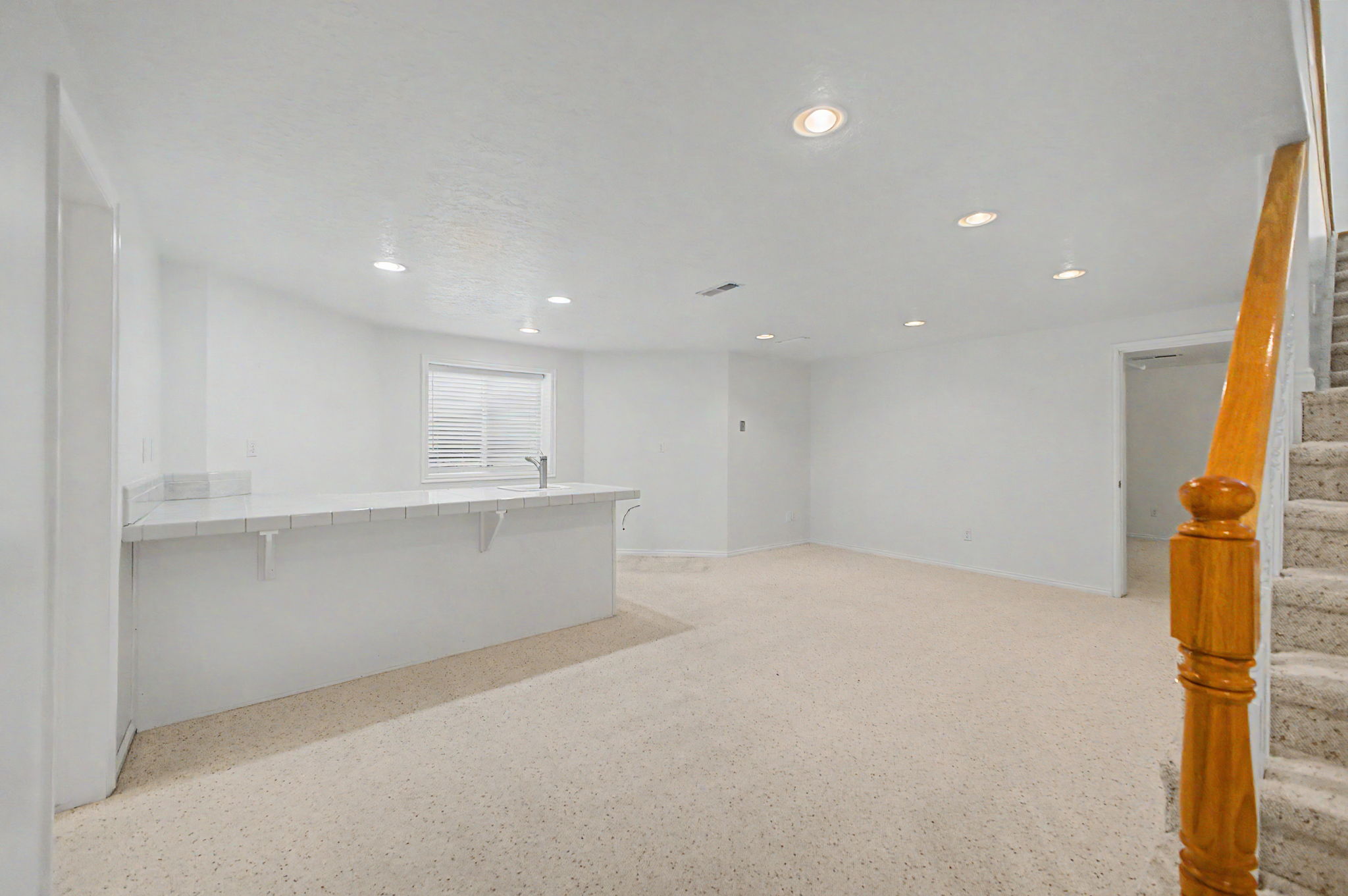 Spacious room featuring a bar with a sink and countertop, recessed lighting, light-colored carpet flooring, a window with blinds, and a wooden handrail beside carpeted stairs