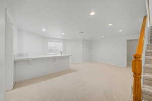 Spacious room featuring a bar with a sink and countertop, recessed lighting, light-colored carpet flooring, a window with blinds, and a wooden handrail beside carpeted stairs