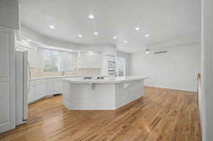 Kitchen featuring a large central island with a cooktop, built-in double ovens, recessed lighting, wood-finish flooring, and white cabinetry