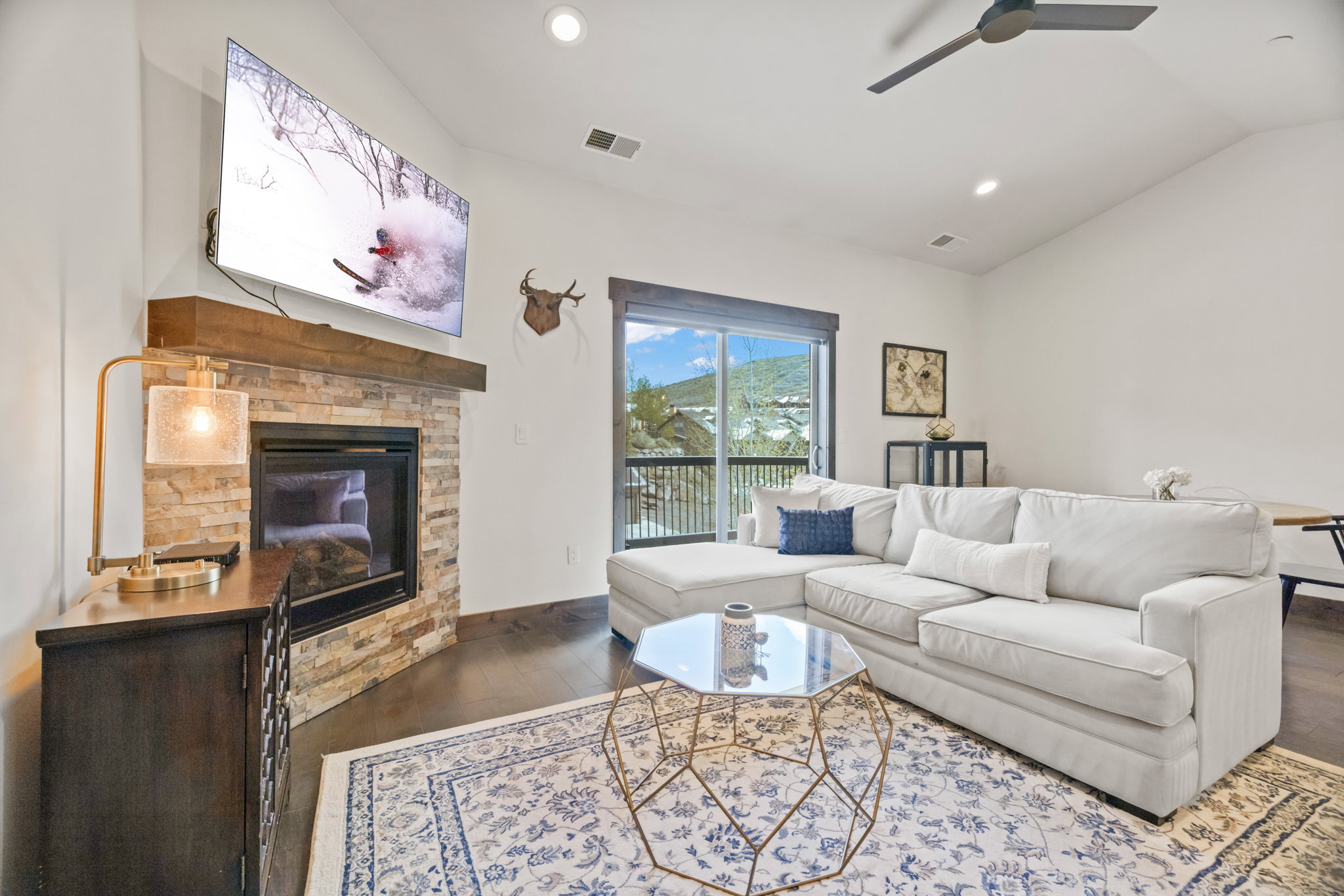 Living room featuring a stacked stone fireplace with a wood mantle, an attached flat-screen television, dark wood-finish flooring, and a sliding glass door opening to an outdoor balcony