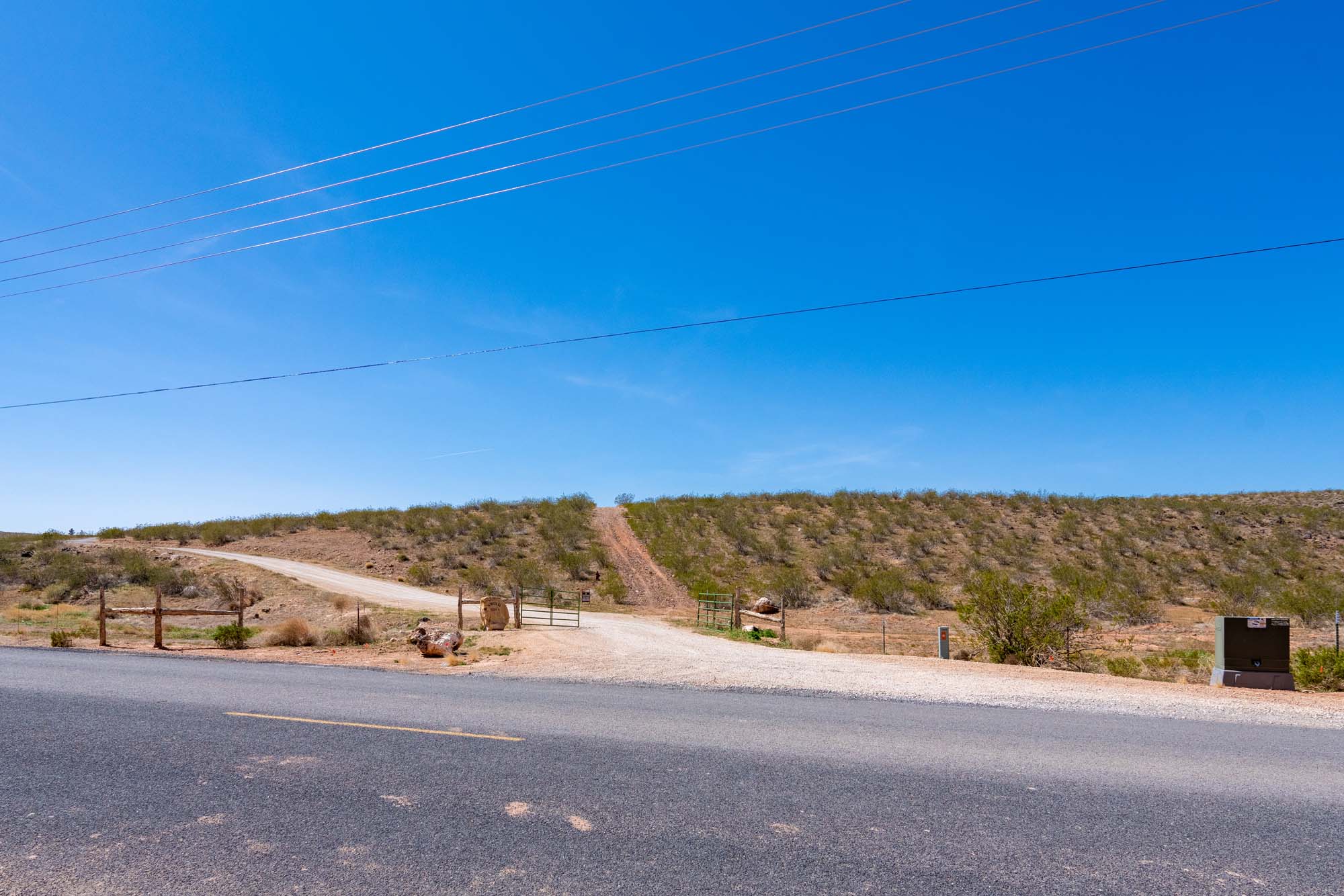 Paved road frontage with a gravel driveway entrance, extending to a fenced gate and an ascending dirt path