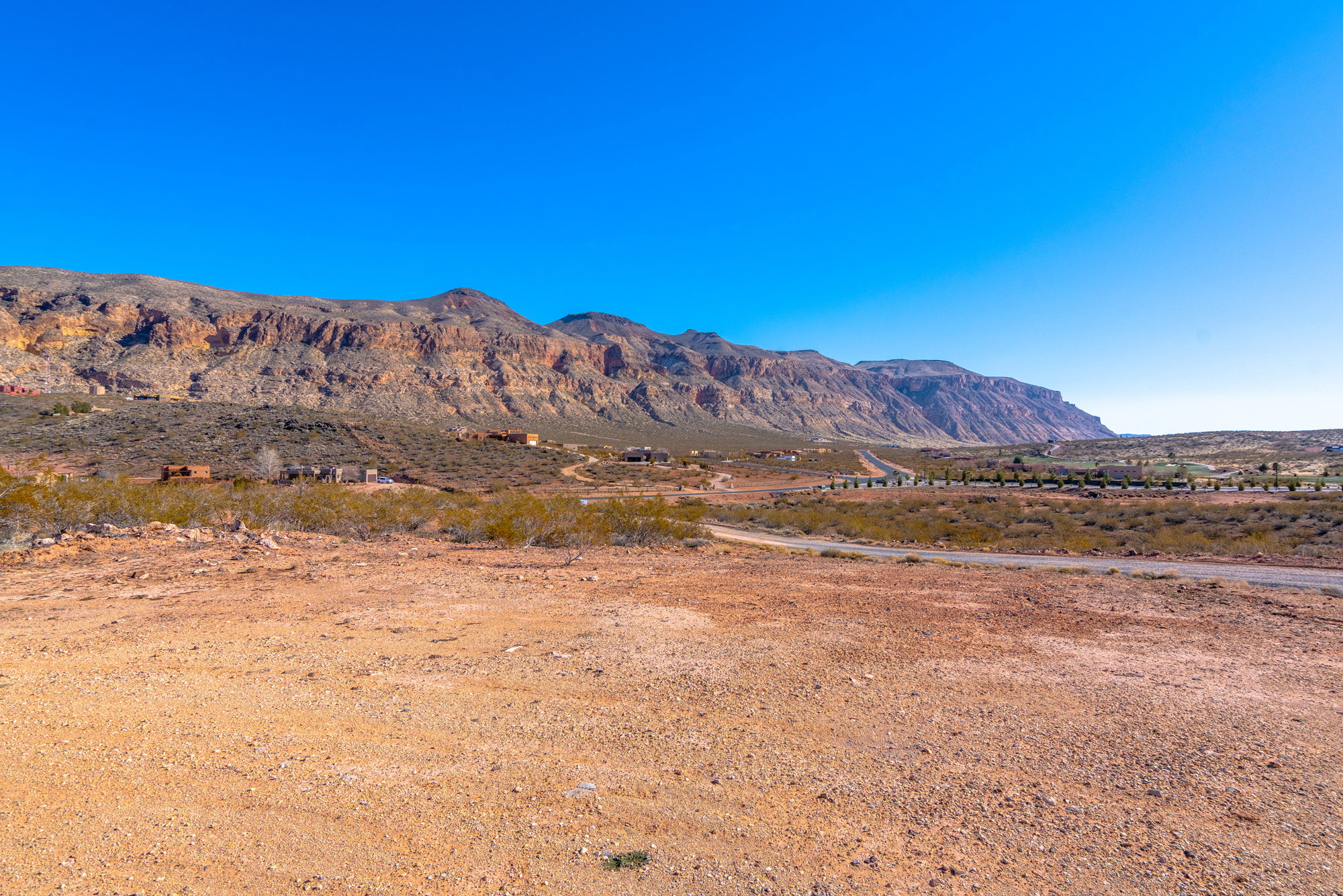 Expansive desert landscape featuring rugged mountain formations, a clear blue sky, distant homes, and a paved road