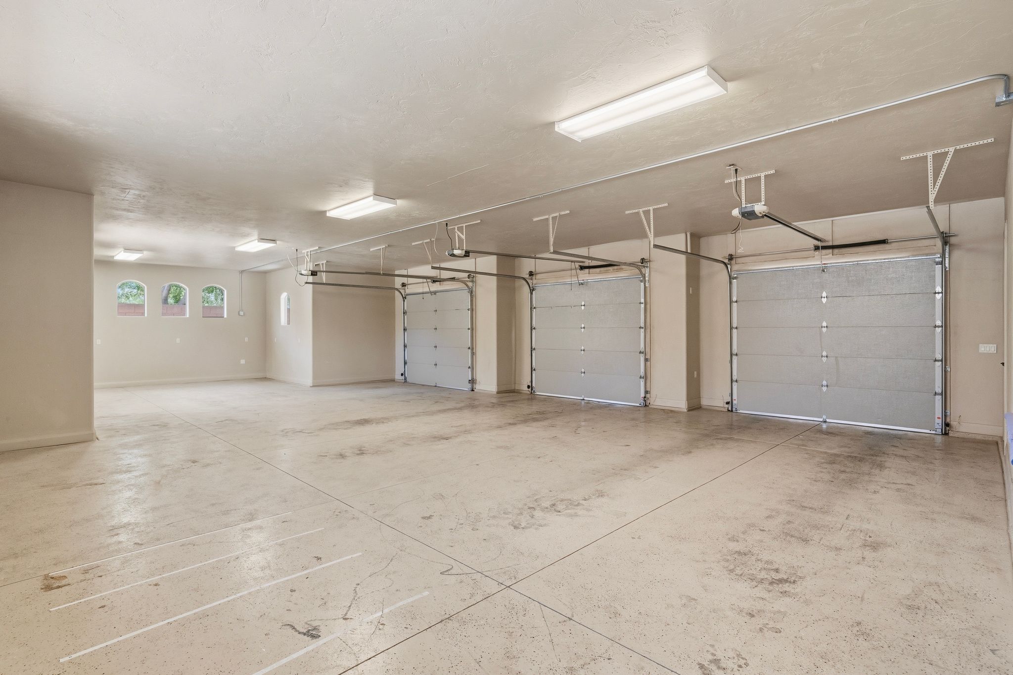 Spacious garage featuring three individual garage doors, concrete flooring, and multiple ceiling-mounted fluorescent light fixtures