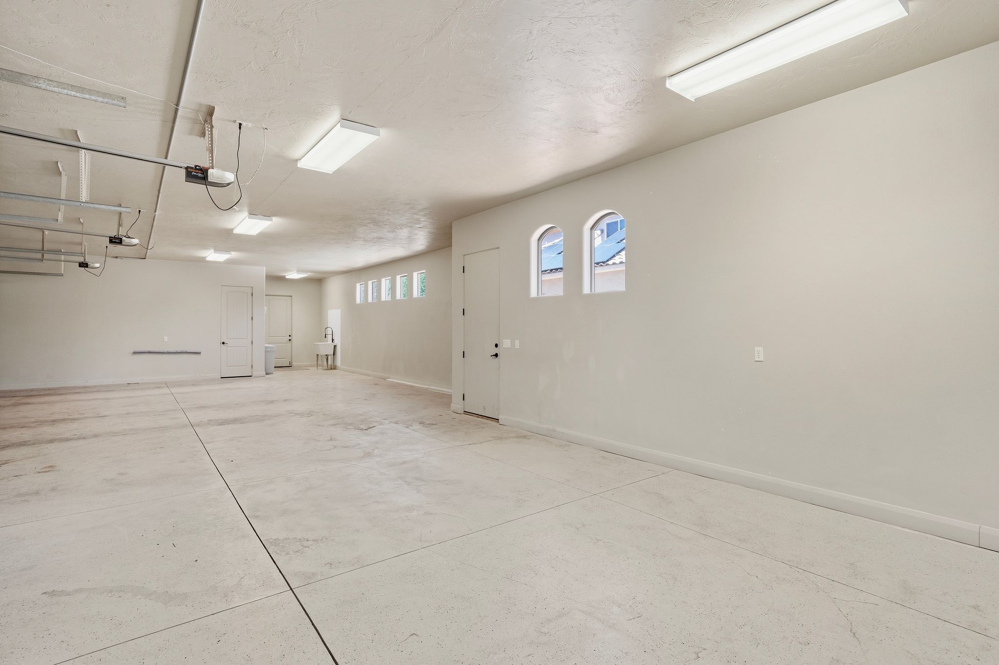 Spacious garage featuring a light-toned concrete floor, multiple fluorescent light fixtures, two white entry doors, and arched windows providing natural light