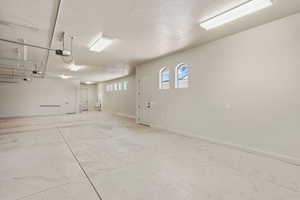 Spacious garage featuring a light-toned concrete floor, multiple fluorescent light fixtures, two white entry doors, and arched windows providing natural light