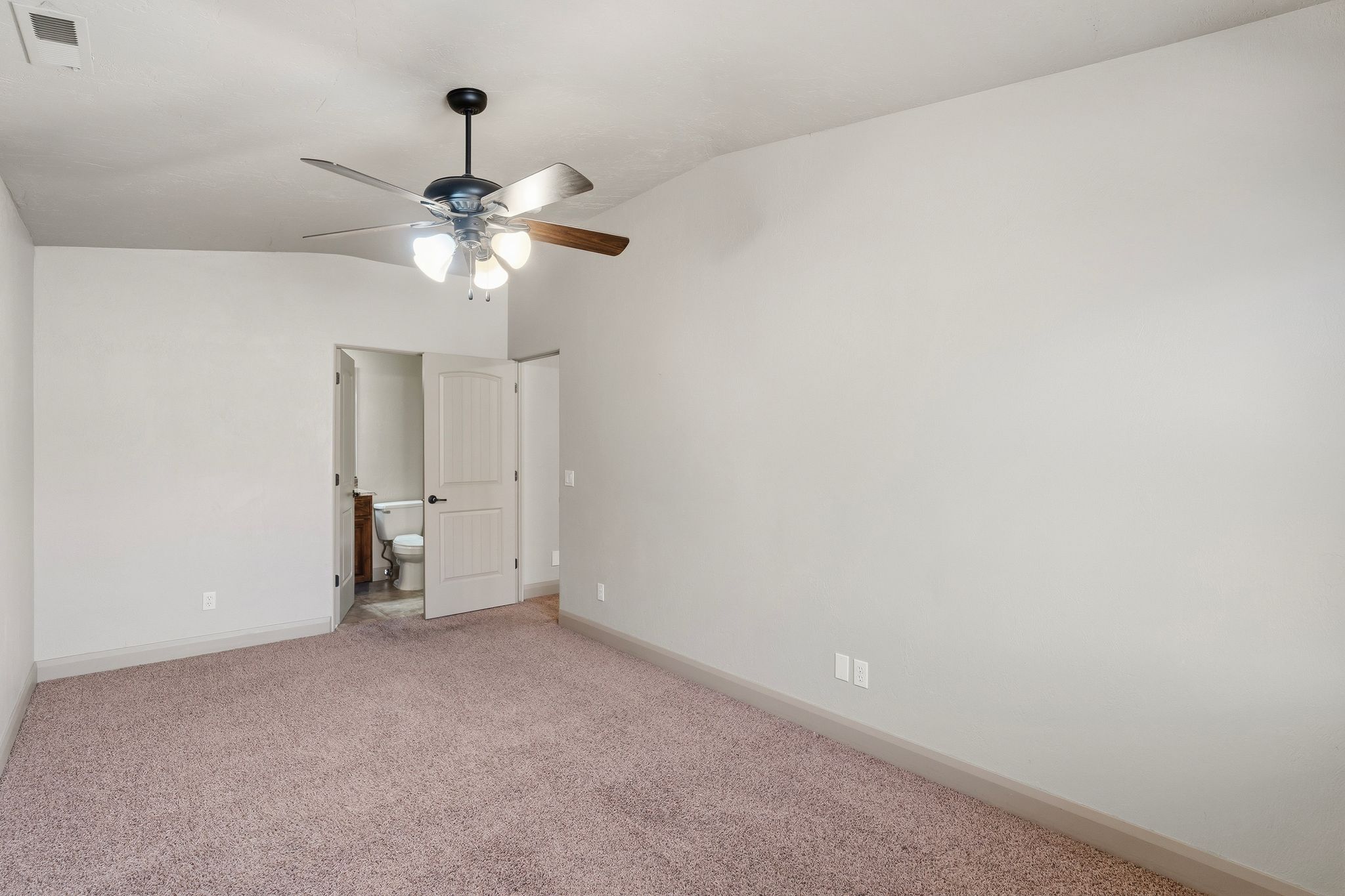 ADU spacious bedroom featuring neutral carpeting, a ceiling fan with integrated lighting, and a white paneled door revealing a bathroom with a toilet and wood-finish vanity