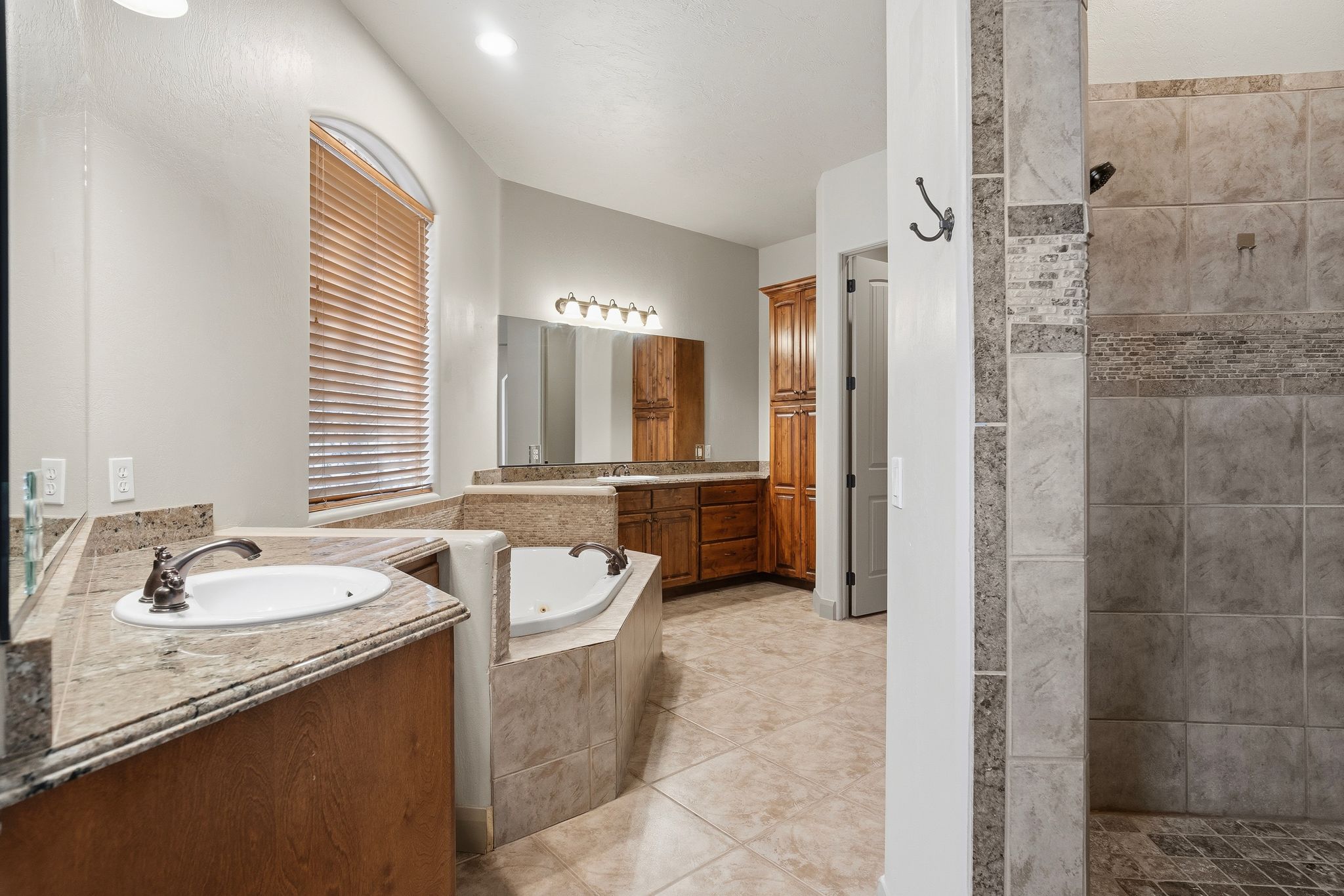 Primary bathroom featuring a built-in tub, a walk-in shower with tile surround, dual vanity sinks with stone-finish countertops, wood cabinetry, and an arched window with blinds