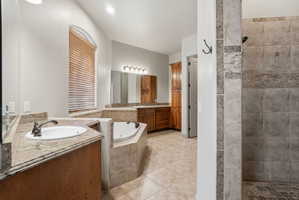 Primary bathroom featuring a built-in tub, a walk-in shower with tile surround, dual vanity sinks with stone-finish countertops, wood cabinetry, and an arched window with blinds