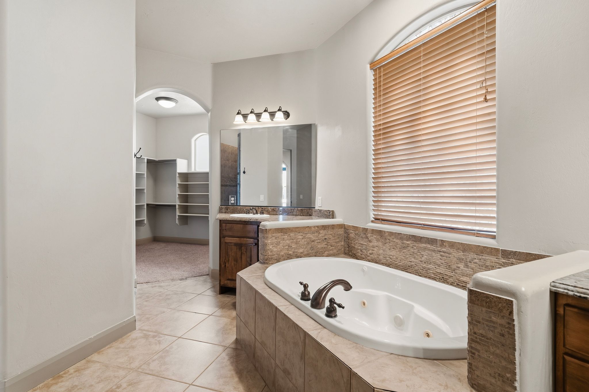 Primary suite bathroom, Jetted soaking tub with tile surround, arched window with horizontal blinds, and a vanity with a rectangular sink, bronze-finish faucet, and a large mirror