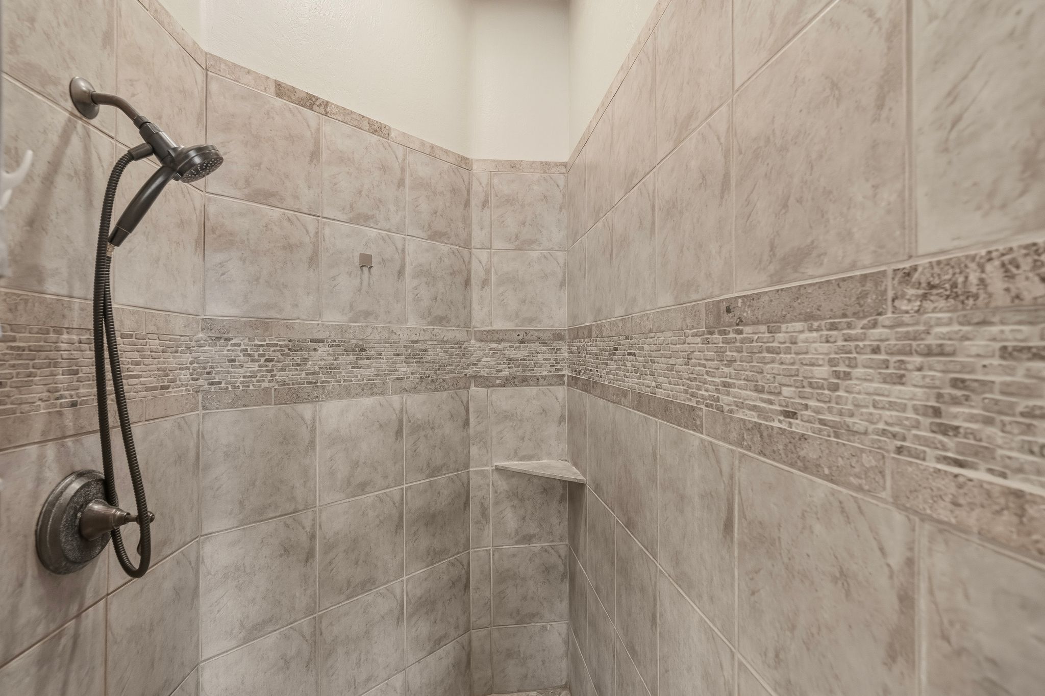 Primary suite bathroom shower enclosure featuring large format tile, a decorative mosaic accent band, a built-in corner shelf, and a bronze-finish shower head with handheld sprayer