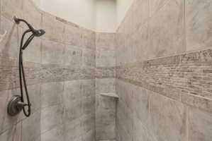 Primary suite bathroom shower enclosure featuring large format tile, a decorative mosaic accent band, a built-in corner shelf, and a bronze-finish shower head with handheld sprayer